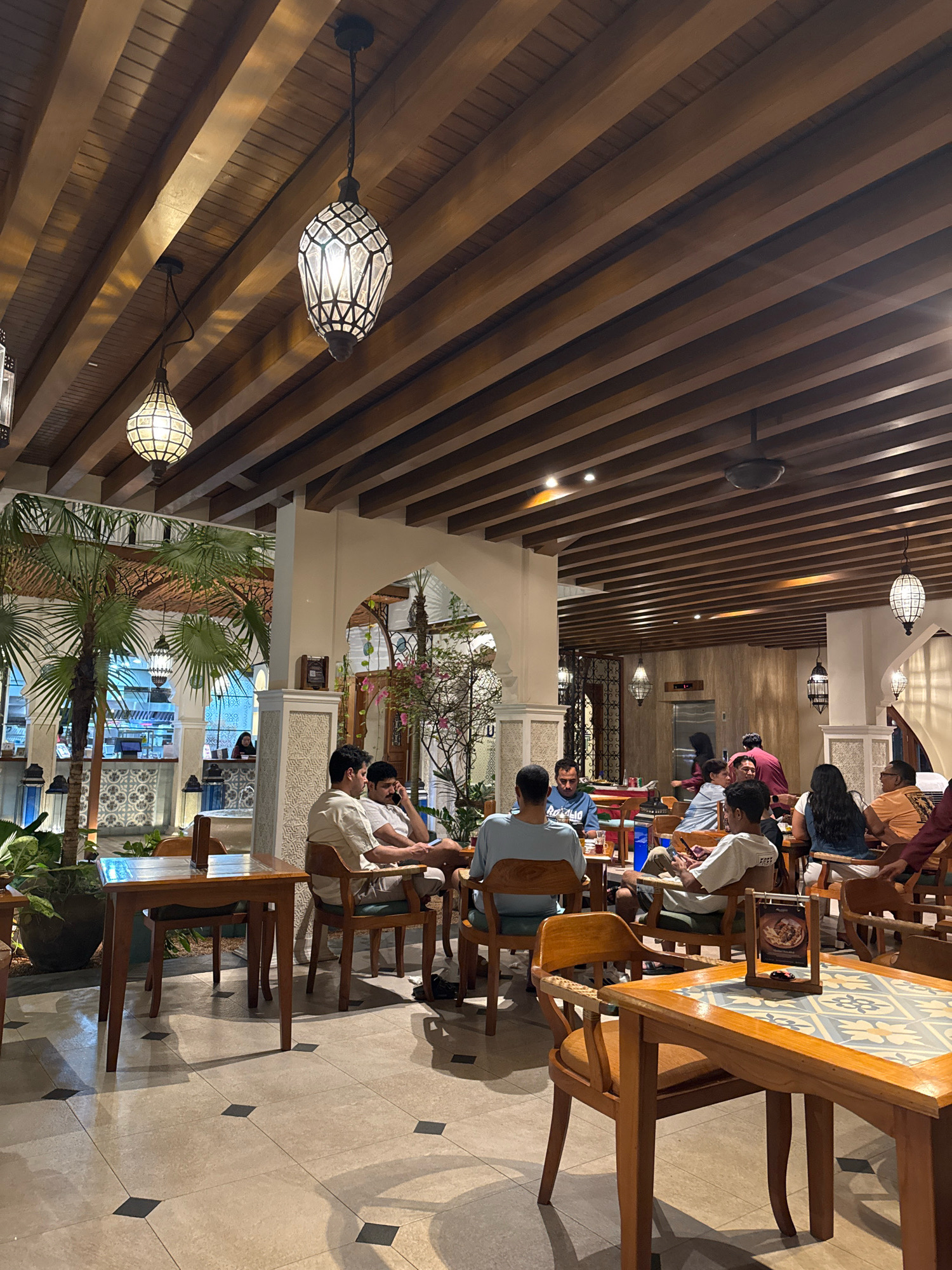 The image depicts the interior of a restaurant in Denpasar, Indonesia. The dining area is illuminated by decorative lanterns hanging from a wooden beam ceiling. Several tables and chairs are occupied by patrons engaged in conversation and dining. In the foreground, a wooden table with a patterned tiled top is visible, with chairs placed around it. In the mid-ground, a group of people are seated at various tables, some interacting with each other and others looking at their phones. A reception desk is visible in the background, with staff attending to guests. Palm trees and potted plants are strategically placed throughout the restaurant, adding to the ambiance. The overall atmosphere appears to be relaxed and social, with customers enjoying their meals in a well-lit and tastefully decorated environment.