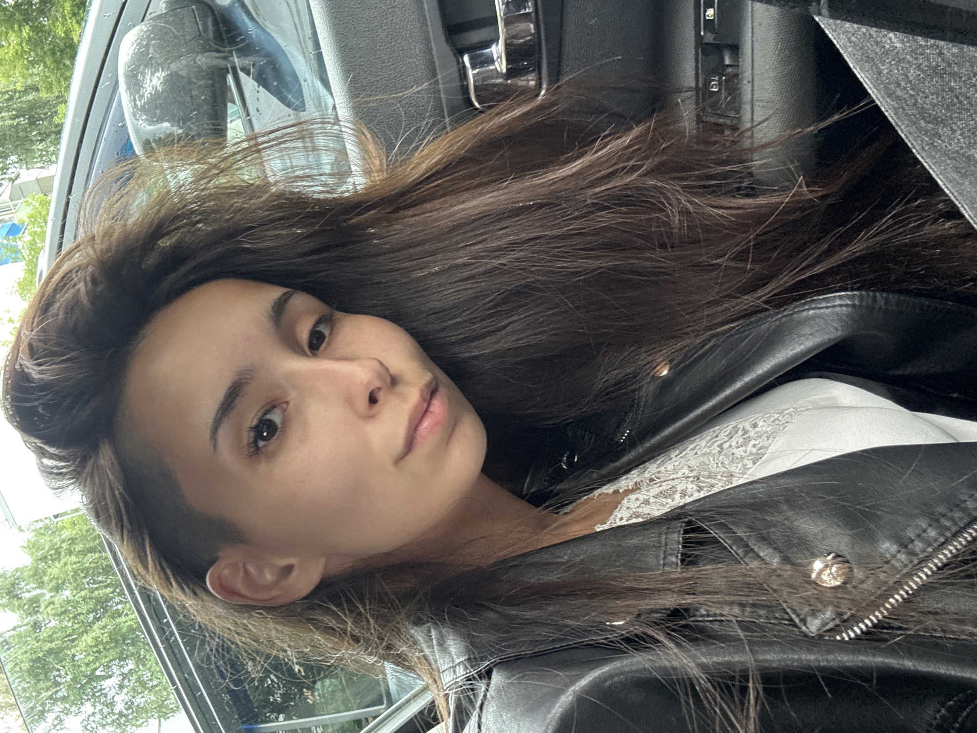 A young woman with long, dark brown hair, styled with an undercut on one side, is sitting in the driver's seat of a car. She is wearing a black leather jacket over a white lace top. Her hair is windswept, partially obscuring her face. The car's interior is visible, including the door panel and a side mirror. Outside the car, trees and a building can be seen, suggesting an urban or suburban setting. The scene appears to be captured in Izhevsk, Russia, likely during daytime, as indicated by the natural lighting.