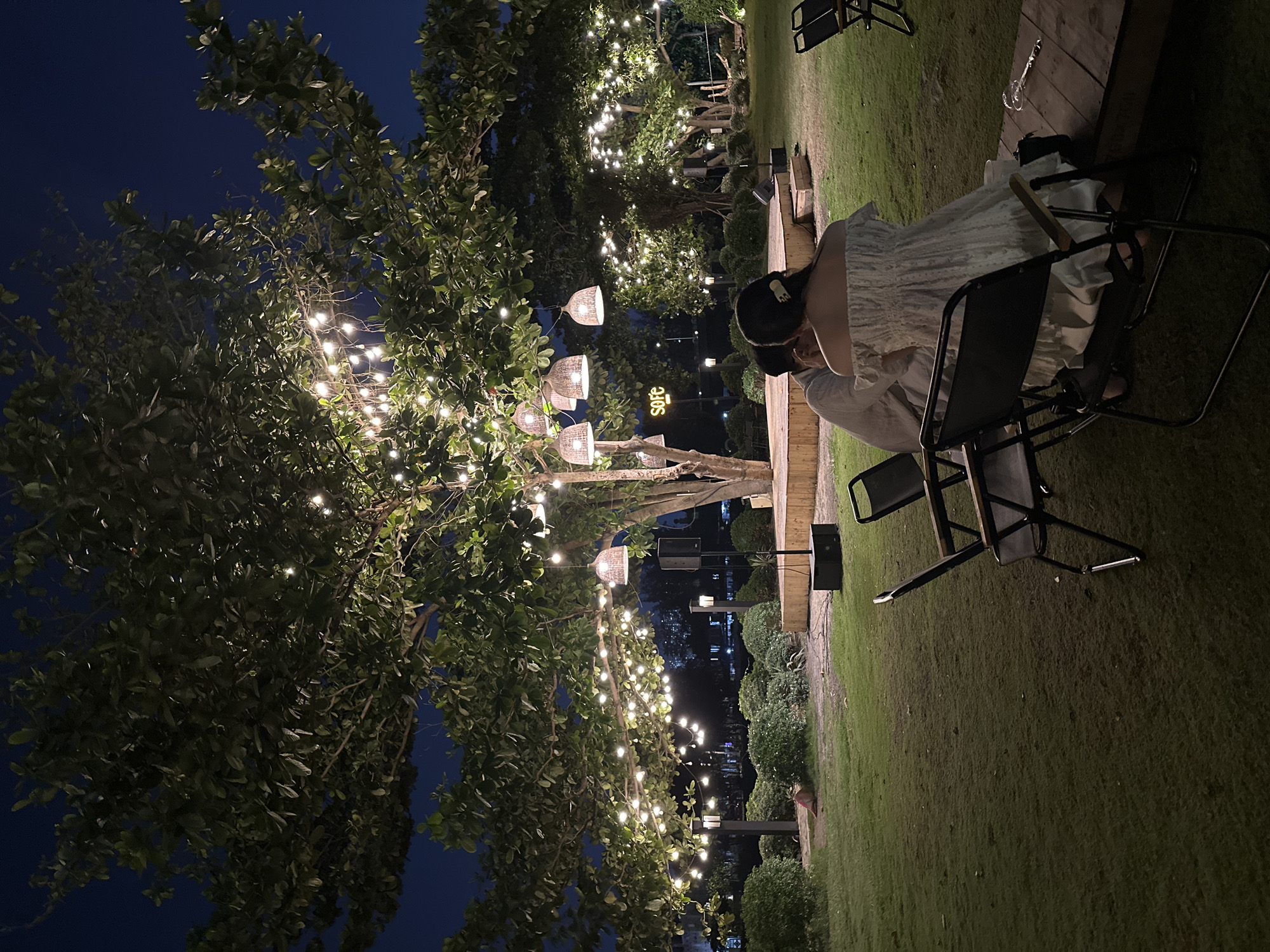 A woman sits in a chair outdoors at night, at an establishment with trees adorned with strings of lights. The location is Ho Chi Minh City, Vietnam. She is wearing a white dress and has dark hair pulled back with a clip. The venue has wooden decking and a lawn area. Other chairs and tables are visible, and the surrounding greenery is lit with more string lights. Buildings can be seen in the distance.