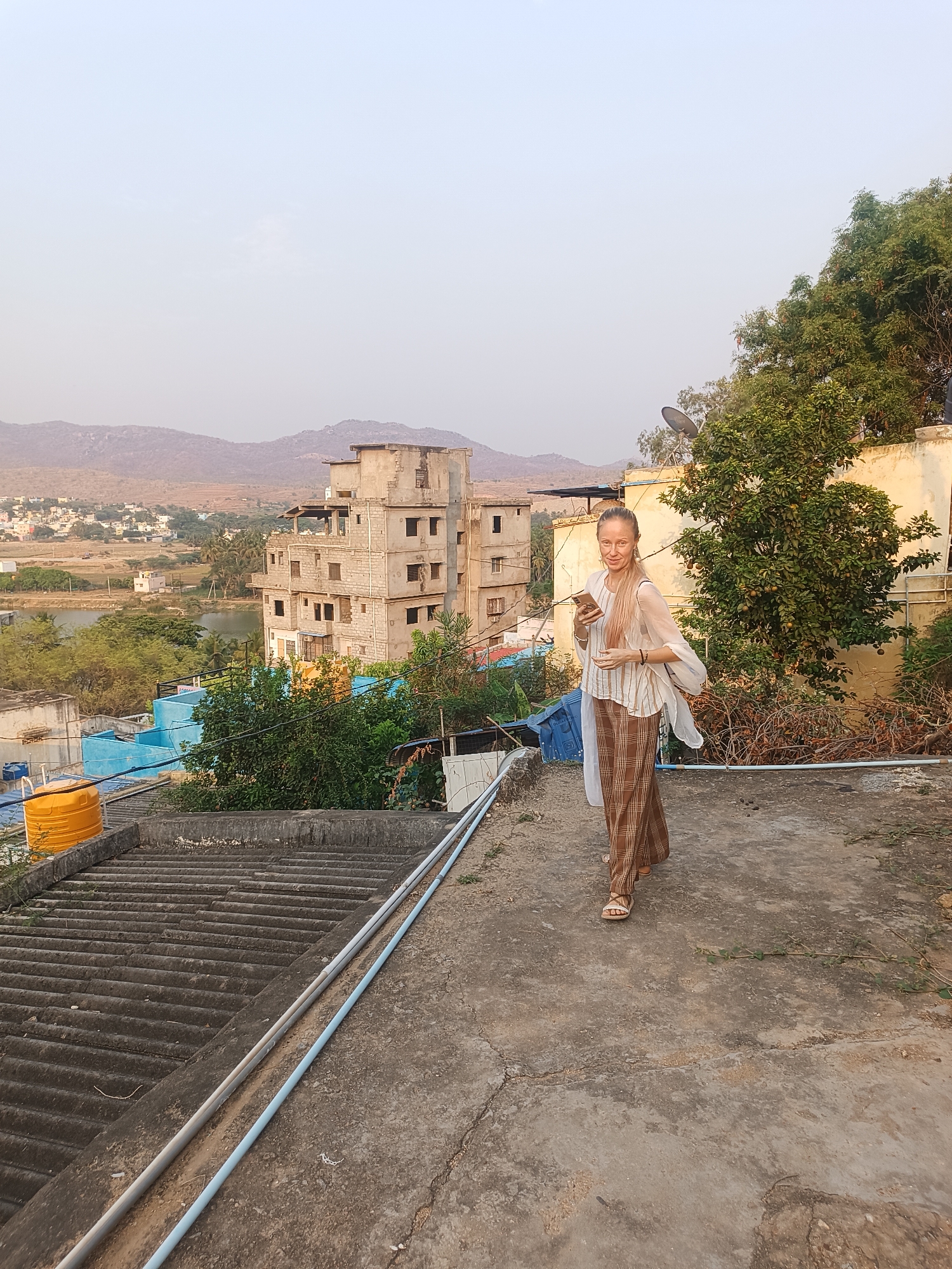 A Caucasian female walks on a concrete rooftop in Puttaparthi, India, holding a smartphone. She wears a light-colored, striped top, brown plaid pants, and sandals. Behind her is a multi-story, unfinished concrete building, a lake, and distant hills under a pale sky. The rooftop is textured with concrete and corrugated metal sheets, featuring exposed pipes and vegetation. A yellow water tank is visible near the lower left corner. A tree and building are to the right of the woman.