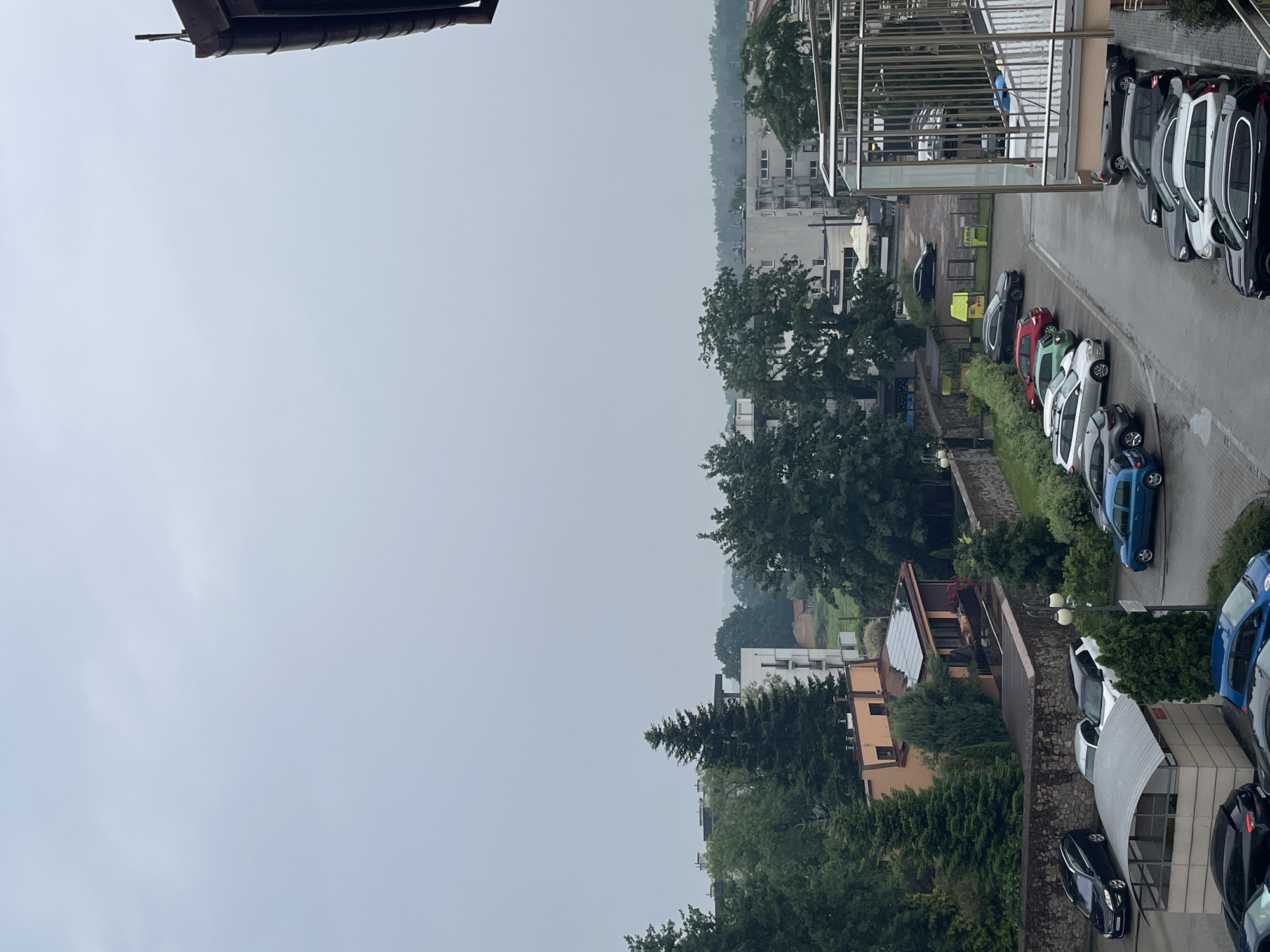 The image shows a view of a street with parked cars and buildings under an overcast sky. The street is wet, suggesting it may have rained recently. Several cars are visible in a parking lot and along the street. Some buildings are multistory apartment buildings, while others appear to be smaller houses or commercial structures. Trees and foliage are present throughout the scene. A metal railing and balcony are visible in the foreground on the right side of the image, indicating this is likely a view from a higher elevation. The overall atmosphere is muted due to the cloudy weather.