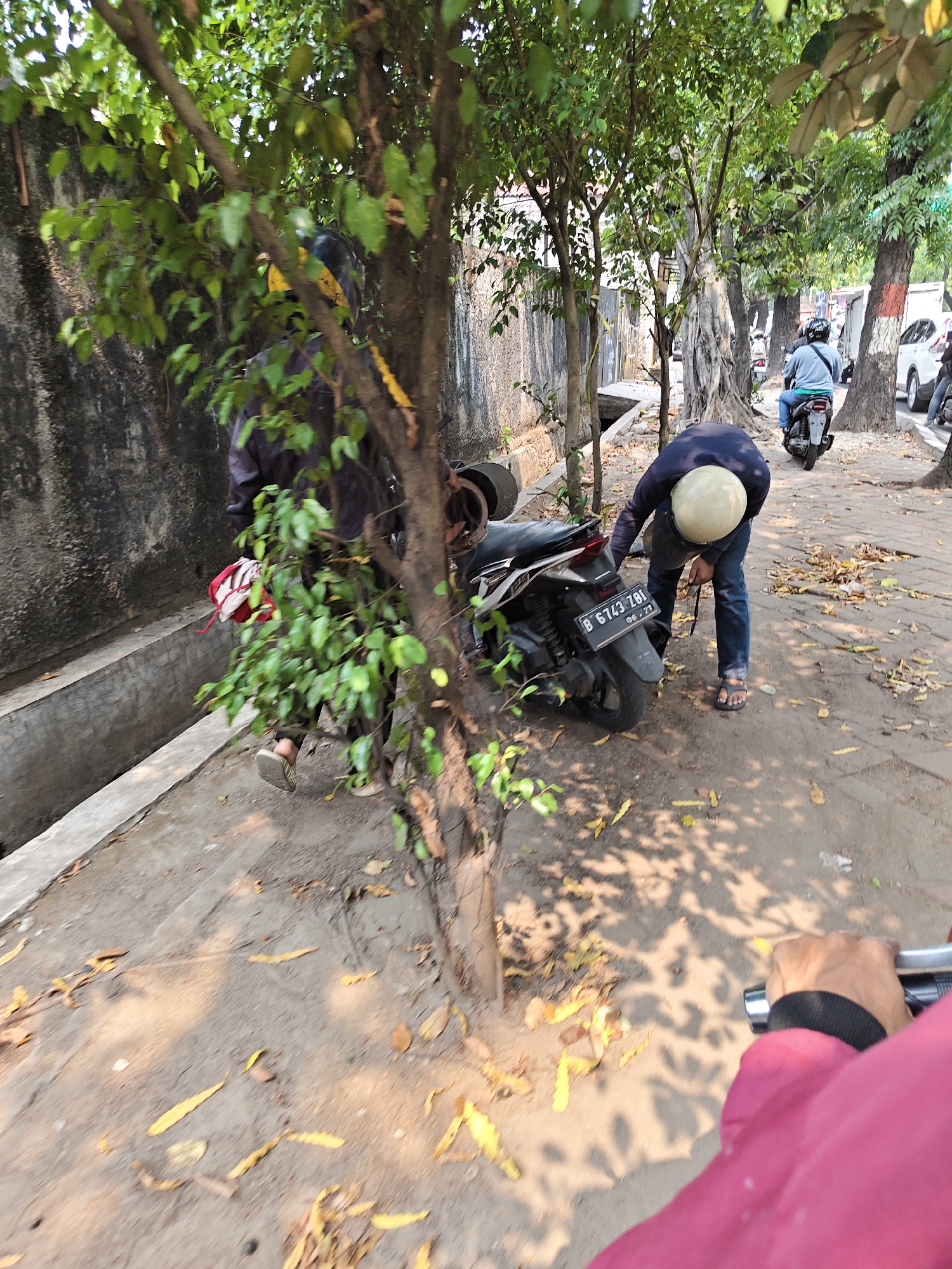 In West Jakarta, Indonesia, a person wearing a navy blue long-sleeved shirt, blue jeans, and sandals is bending over a black and white motorcycle. The motorcycle is parked on a dirt path next to a concrete ditch. The person is wearing a light-colored helmet and appears to be adjusting something on the motorcycle. Another person wearing a dark jacket and a yellow helmet is partially visible behind the motorcycle, near a tree. The scene is outdoors, with trees and sunlight casting shadows on the ground. Fallen leaves are scattered on the path. In the background, other motorcycles and vehicles are visible, suggesting a street or roadside environment.