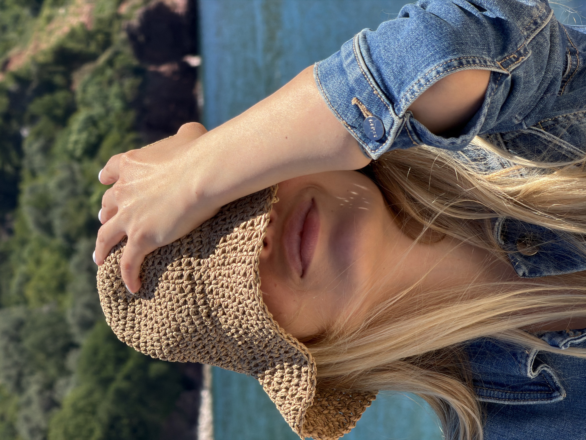 A woman is captured in a medium, vertical shot against an out-of-focus background featuring a blurred landscape of green trees and blue water. She is facing the camera at an angle and using her left hand to hold the brim of a woven sun hat that covers her eyes. Her blonde hair is visible, and she is wearing a blue denim jacket. The location is likely Sveti Stefan, Montenegro, based on the typical landscape elements.