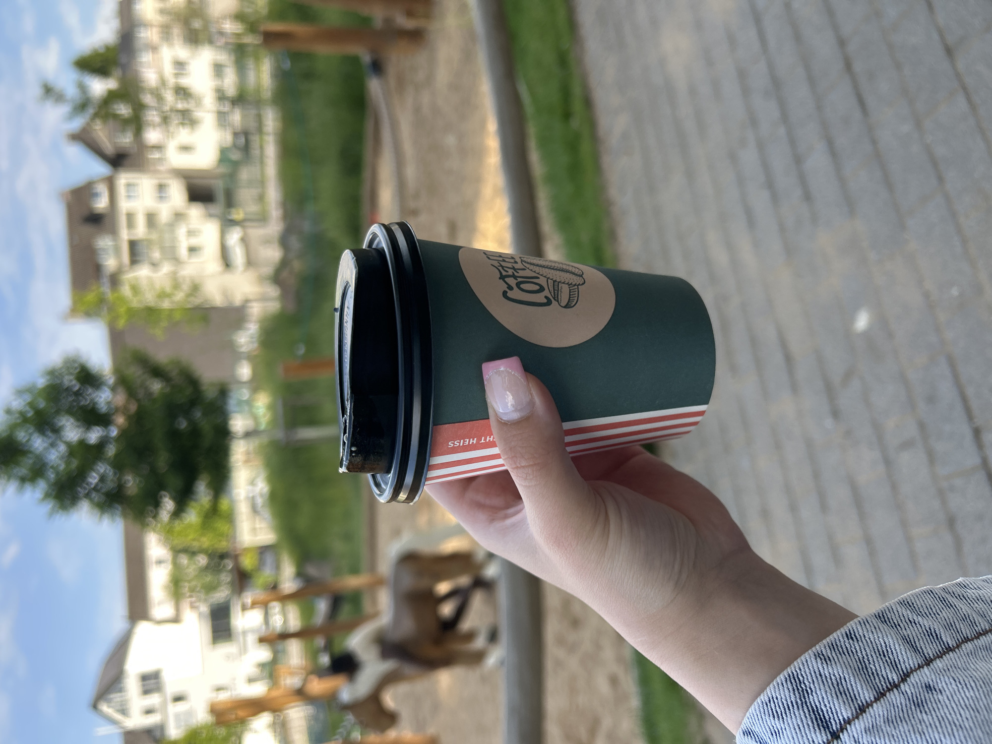In Waldbröl, Germany, a person's hand holds a dark green disposable coffee cup with a black lid. The cup has a label with red and white stripes and a circular brown sticker featuring a coffee bean illustration and the word "Coffee" in a script font. In the background, a playground with a sand pit and wooden structures is visible, including a toy horse. Residential buildings and trees line the horizon under a partly cloudy sky. The ground in the foreground is paved with grey rectangular stones.