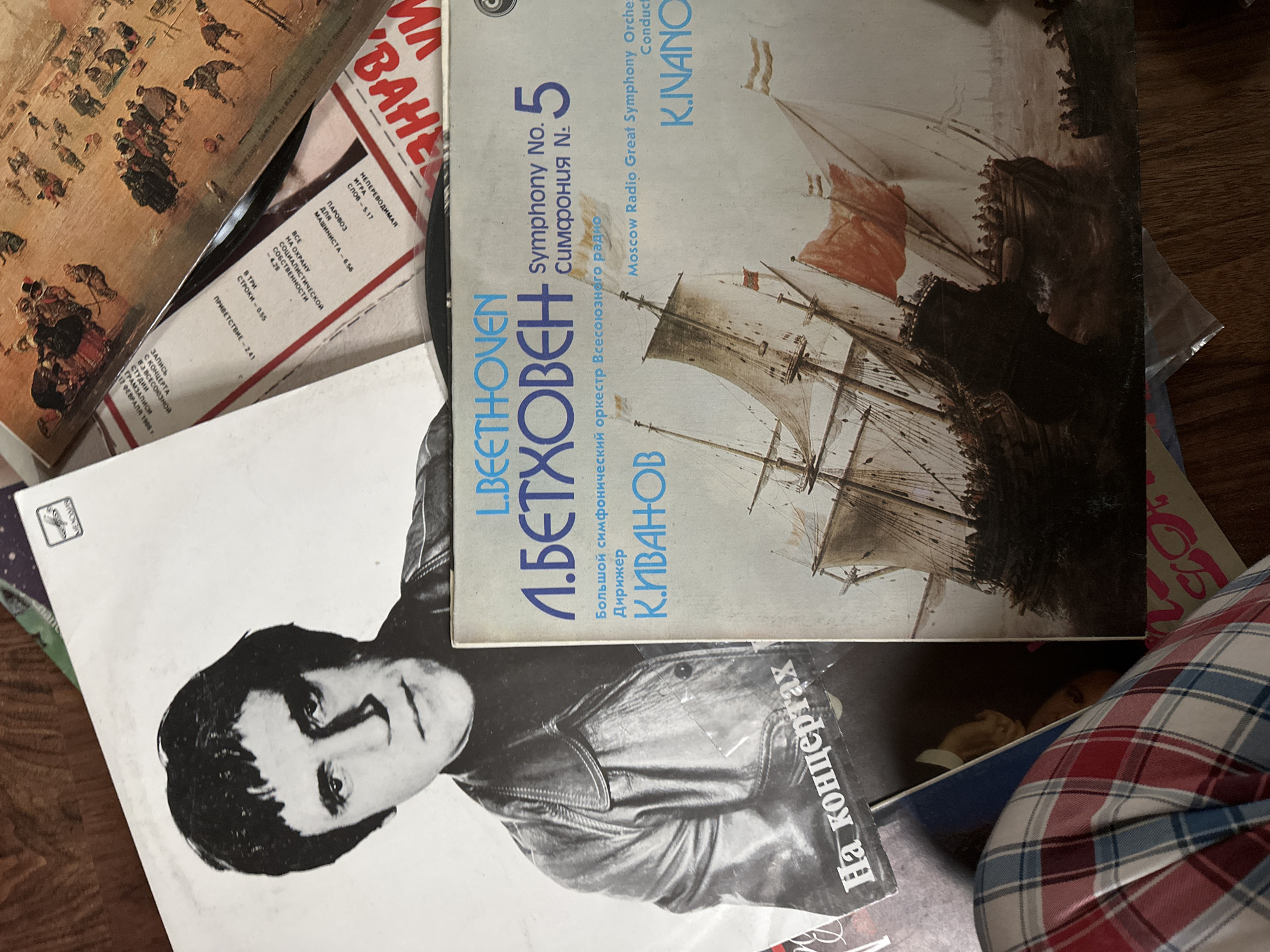 A collection of vinyl records are scattered on a surface. One record features a painting of ships at sea with the words "Beethoven Symphony No. 5" in both English and Russian. The name "K. Ivanov" is also visible. Another record shows a black and white portrait of a man. The words "Na koncertakh" in Russian can be seen. A third record has what seems to be an old painting featuring many people in a snowy scenery, and a text with Russian words and numbers. A corner of a plaid fabric can be seen on the lower right side. The scene does not include enough information to be located in Nizhny Novgorod, Russia.