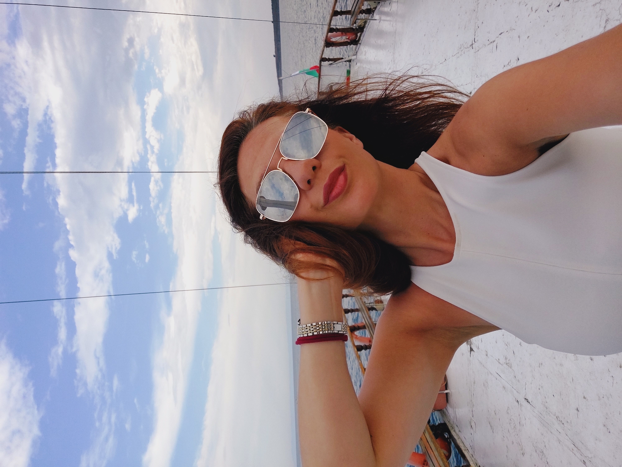 A woman with long, dark brown hair is taking a selfie on the deck of a white boat. She is wearing large, aviator-style sunglasses with reflective lenses and a white sleeveless top. A bracelet made of red cord and silver-colored metallic links is visible on her left wrist. Her left arm is raised, and her hand is resting on the back of her head. The boat's deck is made of white planks. In the background, the sea stretches to the horizon under a partly cloudy blue sky. A wooden railing runs along the edge of the boat. A Bulgarian flag is visible on a pole behind the woman. The scene is likely on the water near Sozopol, Bulgaria, known for its boat tours.