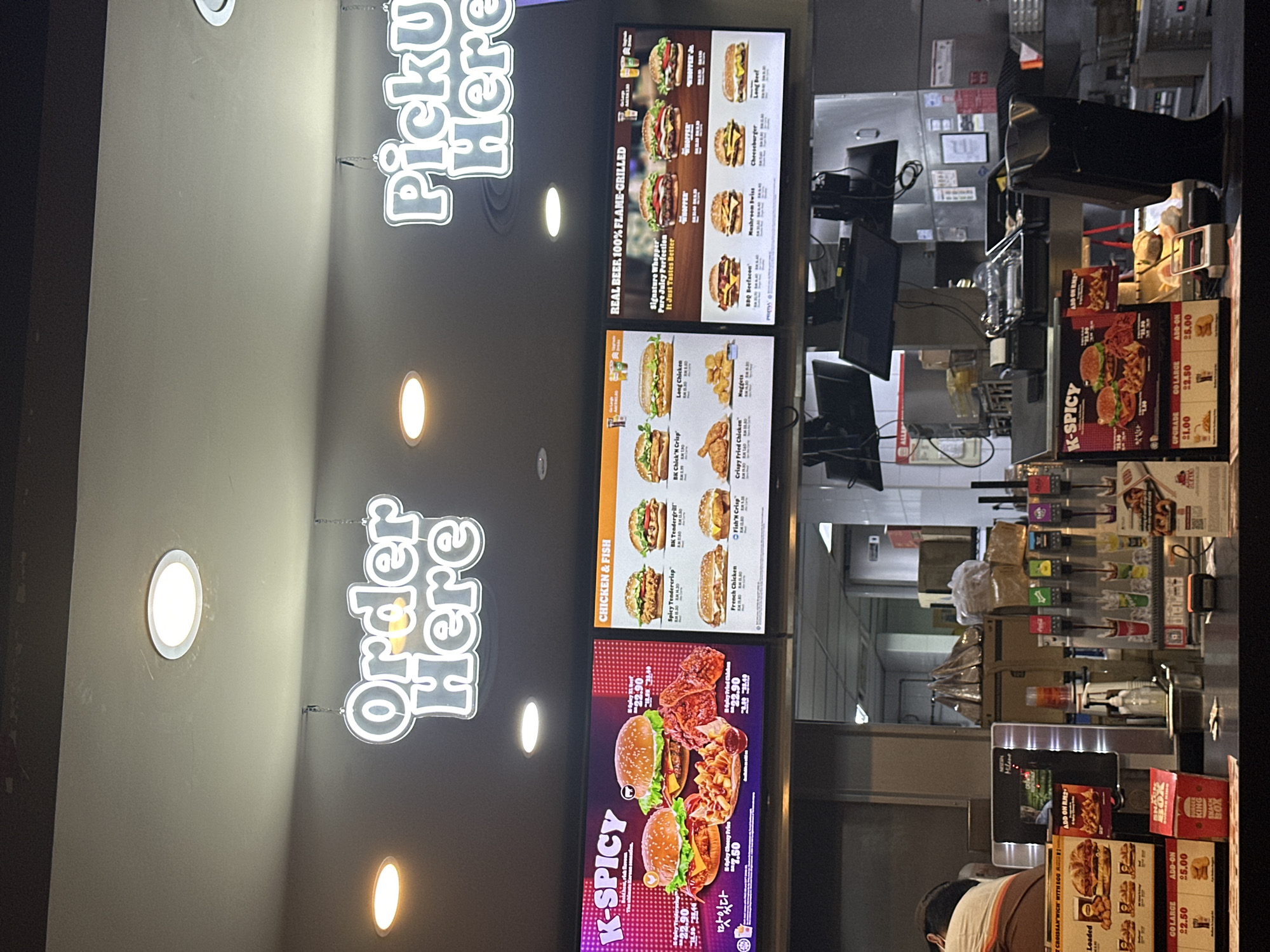 The image depicts the interior of a Burger King restaurant, likely in Subang Jaya, Malaysia, judging by the menu board, which features items such as "K-Spicy" and menu pricing in RM (Ringgit Malaysia). The scene captures a customer placing an order at the counter. The menu boards showcase the "Order Here" and "Pickup Here" signs, along with digital displays advertising various menu items, including burgers, chicken, and fish options. There is a customer, presumably placing an order, visible in the lower portion of the frame. Behind the service counter, drink dispensers, various supplies, and a POS system are visible. The environment is brightly lit.