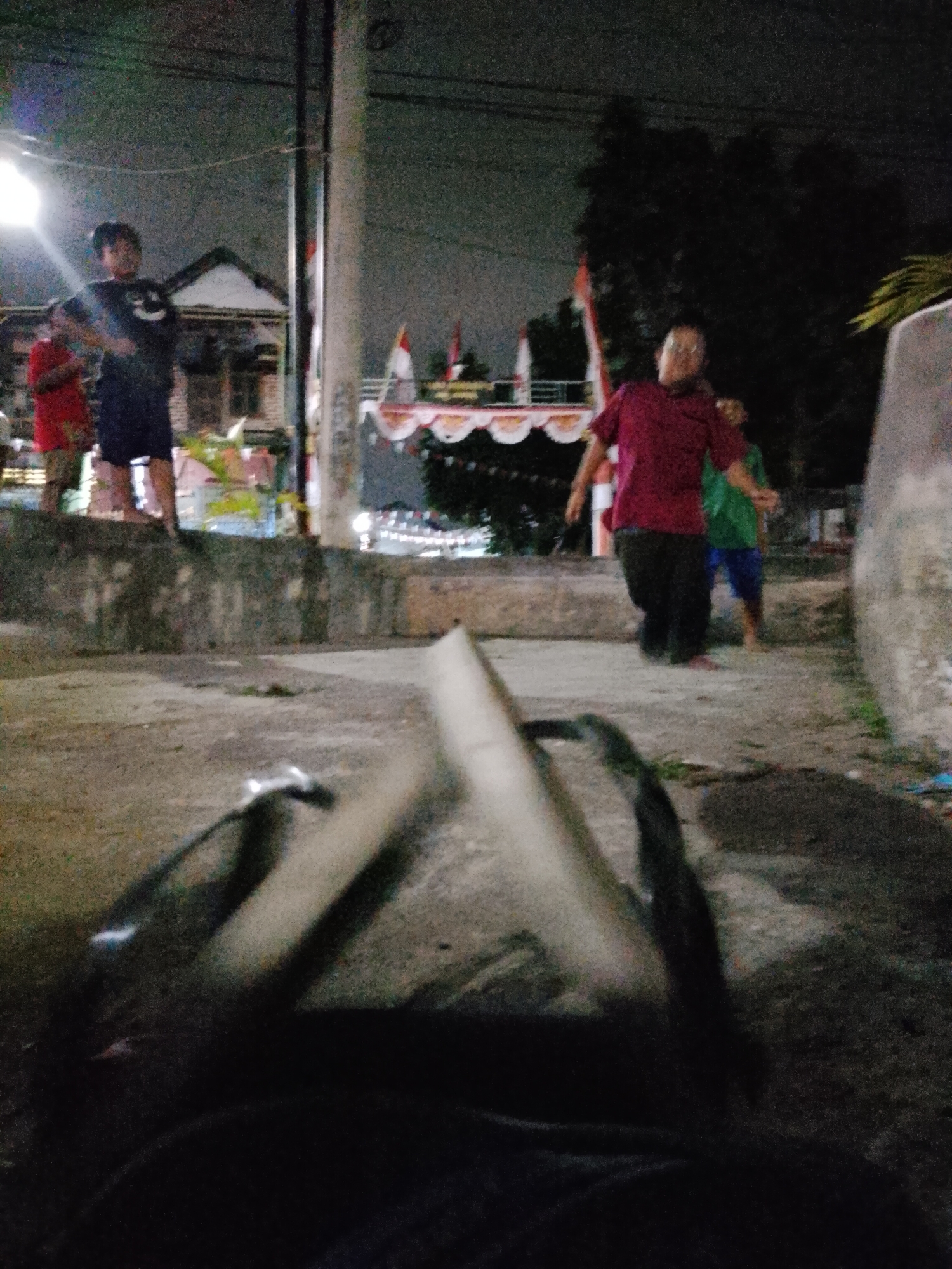 In Pasuruan, Indonesia, at night, a group of individuals are present in an outdoor setting. The foreground is dominated by the blurry depiction of a dark object, possibly a bag or backpack, resting on a concrete surface. Behind this, a group of children are visible. To the left, two boys stand near a concrete ledge. One boy, wearing a black t-shirt with a smiley face graphic, stands beside another boy in a red shirt. The background features houses and a pole with wiring, illuminated by streetlights. In the mid-ground, three individuals are more clearly seen. A man in a maroon shirt and dark pants is captured mid-stride, seemingly running or dancing. To his right, a child in a green shirt and blue shorts is also in motion, possibly playing. Further back, decorations, including flags and bunting, are visible on a building or structure, suggesting a festive occasion. The ground is rough concrete, and the lighting is dim, creating a grainy, low-light effect.