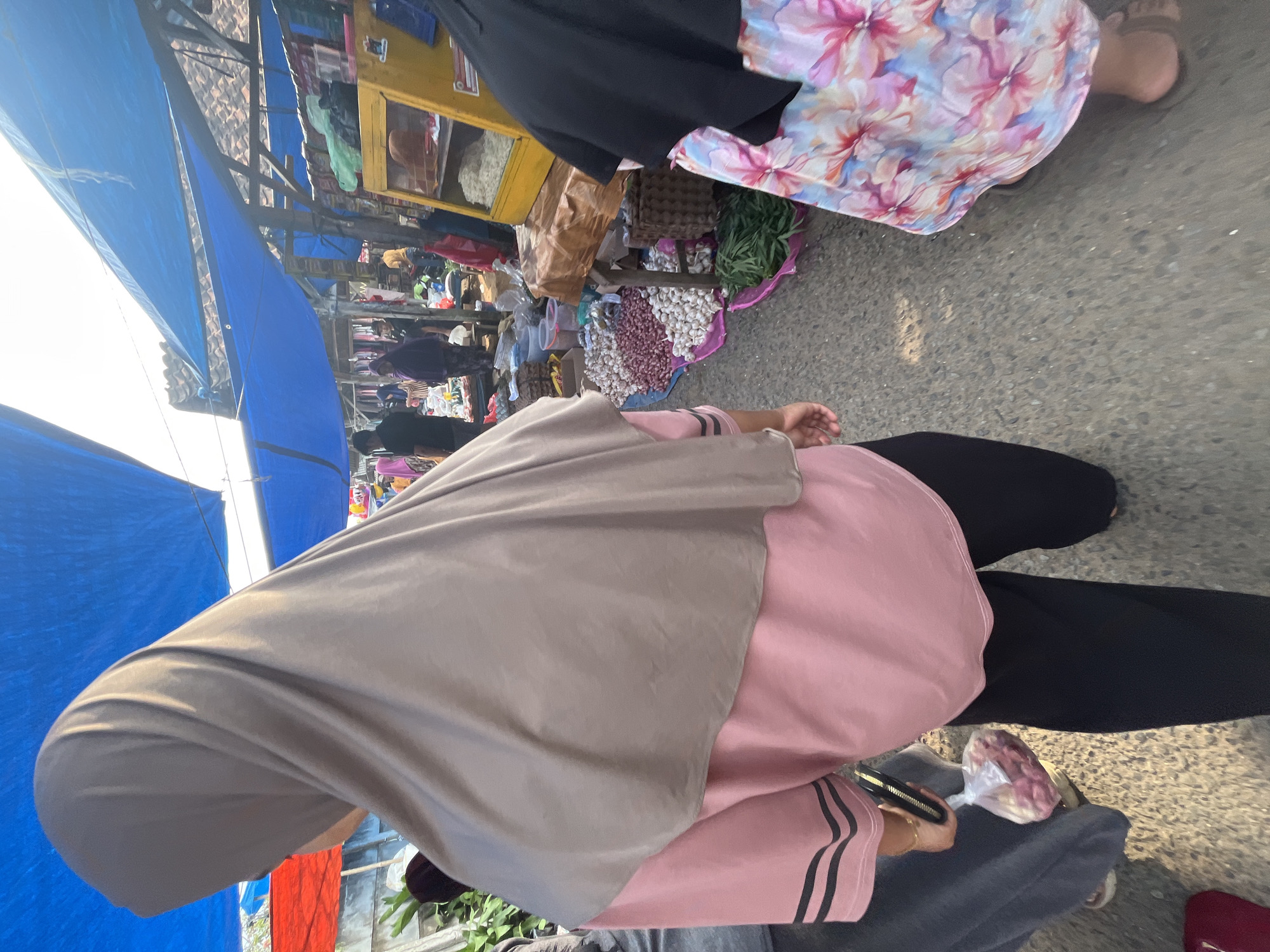 The image depicts an outdoor marketplace scene in Kemuning, Indonesia. In the foreground, a person wearing a taupe hijab and a pink t-shirt with black stripes is seen from behind. They are holding a plastic bag filled with what appear to be red onions and a black wallet. To the right, another person is partially visible, wearing a black hijab and a dress with a floral pattern featuring pink and blue flowers. They are wearing brown sandals.
The marketplace is covered by blue and red tarps stretched over wooden structures, creating a shaded area. Stalls are set up along a paved pathway. One stall displays a variety of produce on the ground, including large piles of red onions and garlic, and bundles of green leafy vegetables. Behind this stall, a yellow wooden display cabinet with shelves holds various items, some of which appear to be food products. Other people are visible in the background, browsing or working at the stalls. The overall atmosphere suggests a busy local market.