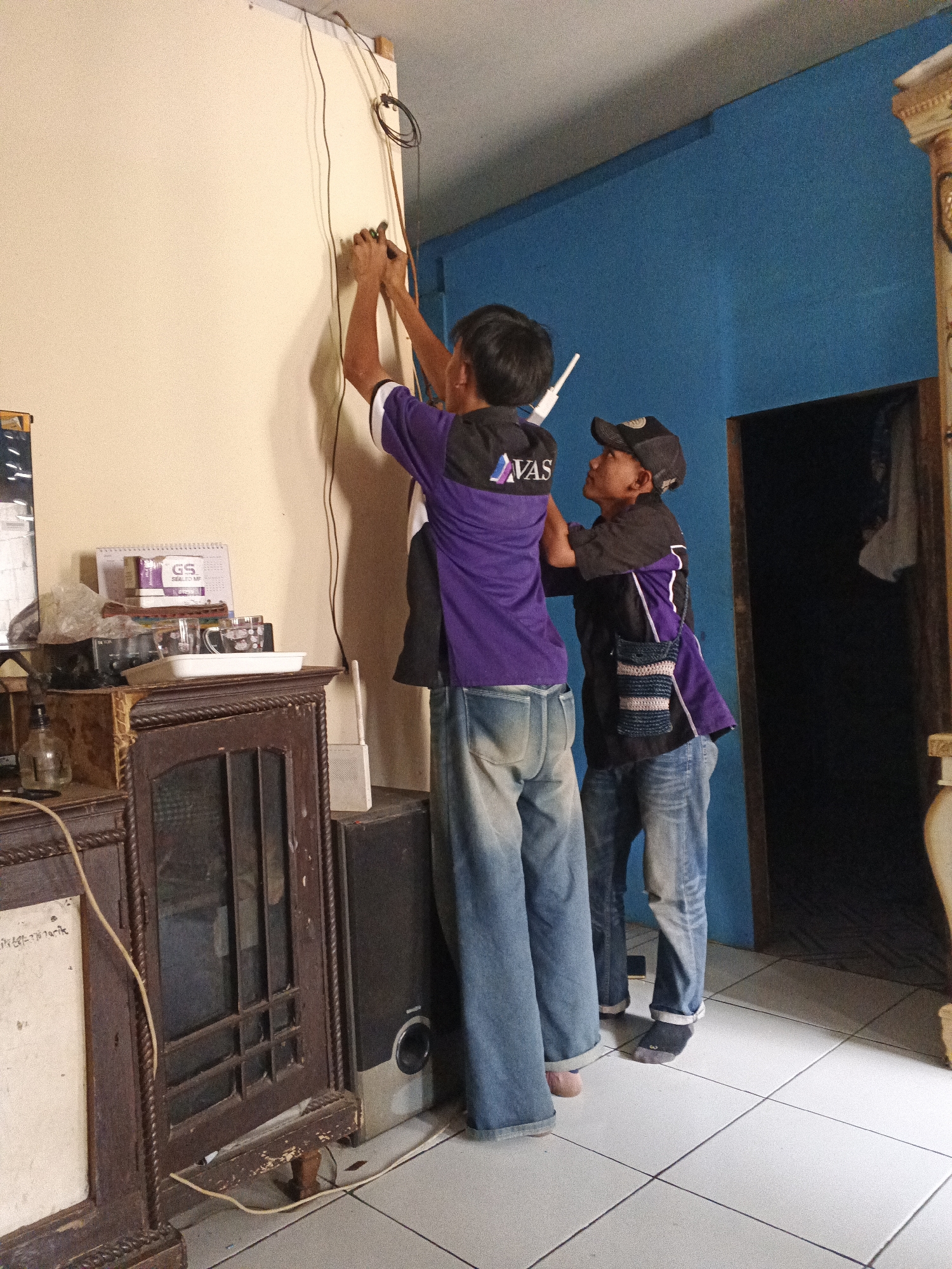 Two young men, likely technicians, are working near a wall in what appears to be a residential room. One man, wearing a purple and black shirt and blue jeans, is standing on the right side of the frame, reaching up to the wall with his right hand, holding what looks like pliers or a similar tool. He is facing away from the camera. The second man, wearing a black t-shirt with a purple accent, jeans, and a black baseball cap, is standing to the left of the first man, also facing the wall. He appears to be assisting or observing. Wires are visible running along the wall and ceiling. In the foreground, to the left, is a dark wooden cabinet with glass doors and some items on top, including a calendar and cups. To the right of the cabinet, a black speaker is visible. The room has light-colored tiled flooring and a bright blue wall on the right side. The overall setting suggests an indoor task related to electrical or network installations. The image was taken in Warungkiara, Indonesia.