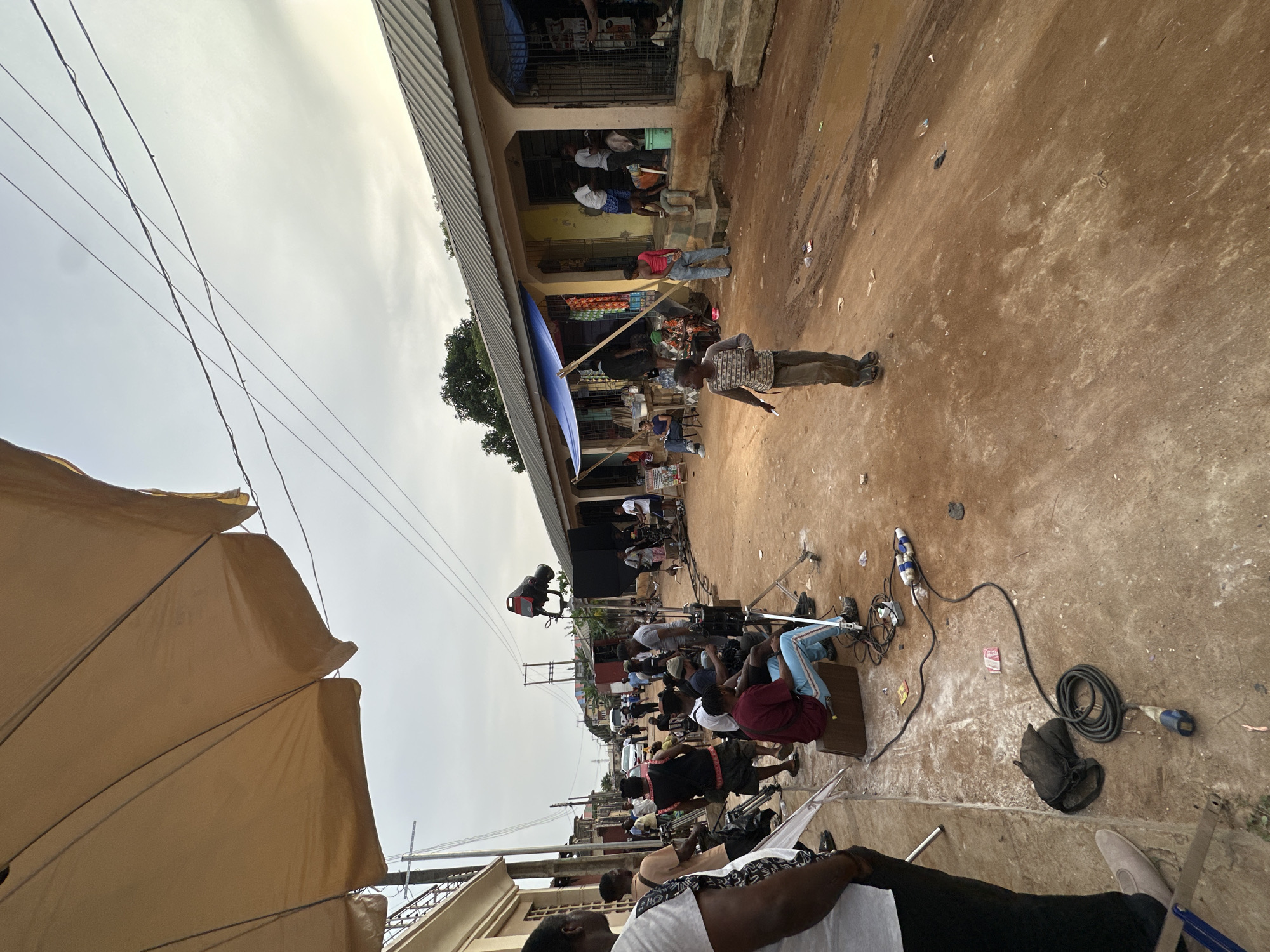 An outdoor film or video production is captured on a reddish-brown dirt surface, bustling with people and equipment. The scene is bordered by low-rise buildings on the right and an open, cloudy, or hazy sky overhead.
In the foreground, a large, light-colored umbrella or awning partially obscures the left side of the frame, with utility lines stretching across the sky. The unpaved ground is composed of reddish-brown earth, scattered with some litter.
Along the right side, simple, low-rise buildings with corrugated metal roofs line the area. Several individuals are seen standing or sitting under the buildings' overhangs, some near what appear to be storefronts. One section of the building features a blue tarp extended as an awning.
The mid-ground is a hive of activity, populated by film crew members. Many individuals, primarily men, are seated on small stools or chairs, facing the production area. Visible equipment includes tripods, potentially holding cameras or lighting fixtures, and black speaker-like boxes or monitors on stands. A prominent light fixture on a tall stand is positioned further down the path. Numerous black cables and power strips are spread across the dirt, with one cable neatly coiled. A man in a patterned shirt and light trousers stands centrally, observing the scene.
The image depicts a dynamic film set in a community environment, characteristic of Lagos, Nigeria. The time of day appears to be daytime, under a diffused or overcast sky. No discernible text is visible.