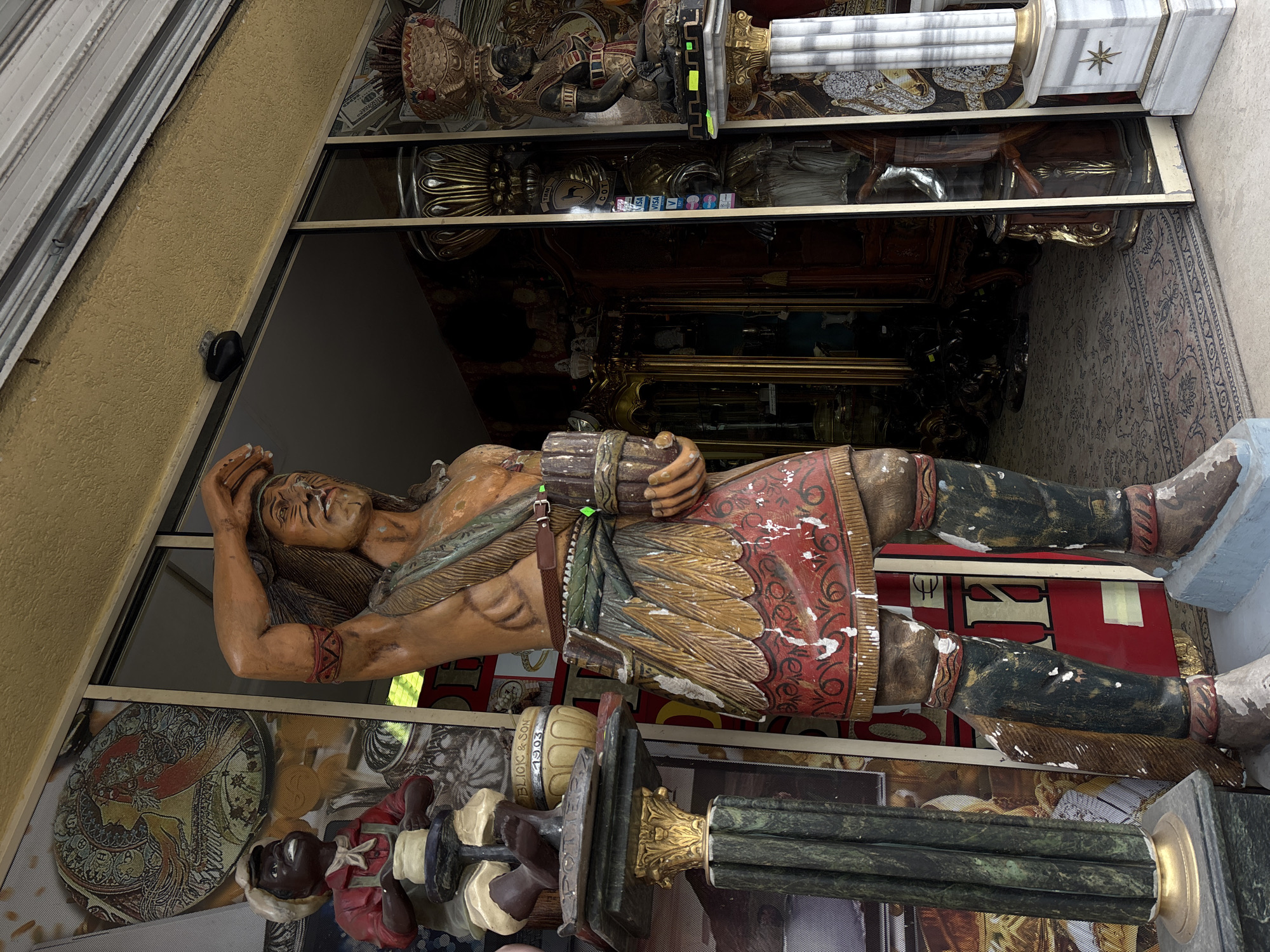 The image captures a static display within what appears to be a shop window in Varna, Bulgaria, showcasing a collection of statues and ornate objects.
The central subject is a large, intricately carved wooden statue of a Native American man, possibly a cigar store Indian. He stands tall, with dark hair, a headband, and painted facial features. His right hand is raised to his forehead, while his left arm cradles a bundle. He is adorned in a feathered kilt or skirt in red, gold, and green, along with armbands and dark boots, all showing signs of age and chipped paint.
Behind the main statue, viewed through the display glass, is a rich assortment of items. These include golden pedestals, a fluted marble column with a star design, and another smaller statue of a dark-skinned figure, resembling a jockey, holding a spool. The base of this smaller statue bears the text "Blick & Son 1903." The interior of the shop visible through the display appears to be darkly lit, containing what look like antique furniture and a patterned rug on the floor. Reflections on the glass overlay the scene, revealing images of US dollar bills and other patterns. A sticker inside the display shows the words "VISA" and "C.O.D."
The scene is illuminated by daylight, suggesting the photograph was taken during the day. No active event is occurring; it is a snapshot of an elaborate storefront display, likely an antique shop or a store specializing in decorative items.