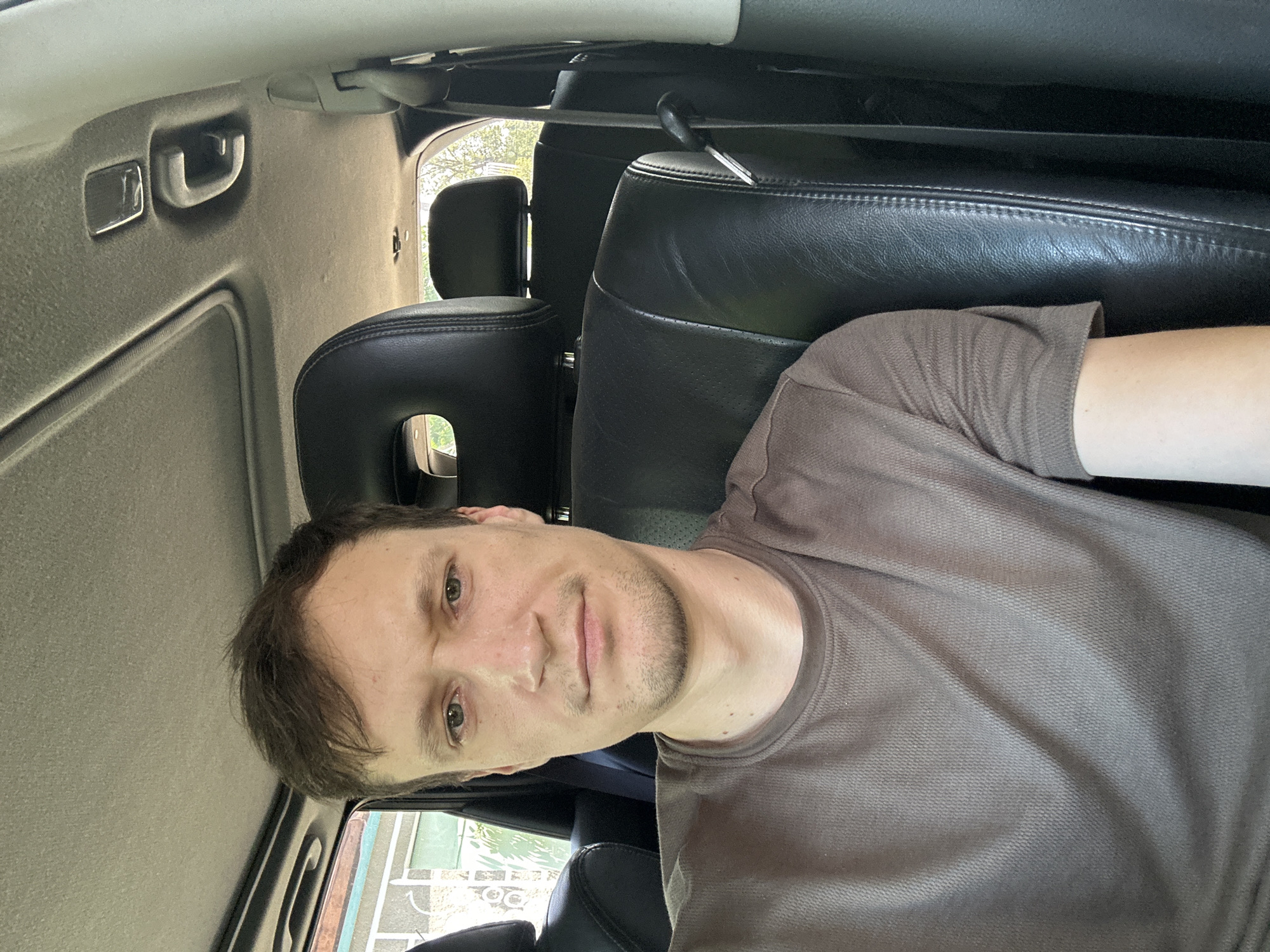 A man with brown hair, a light complexion, and a short beard is seated in the driver's seat of a vehicle. He is wearing a brown t-shirt. The interior of the vehicle is visible, including black leather seats, the ceiling with a sunroof, and a grab handle. Sunlight streams in through the windows, illuminating the scene. The background outside the vehicle shows greenery and a building, suggesting an outdoor location, possibly in the city of Makiv, Ukraine.