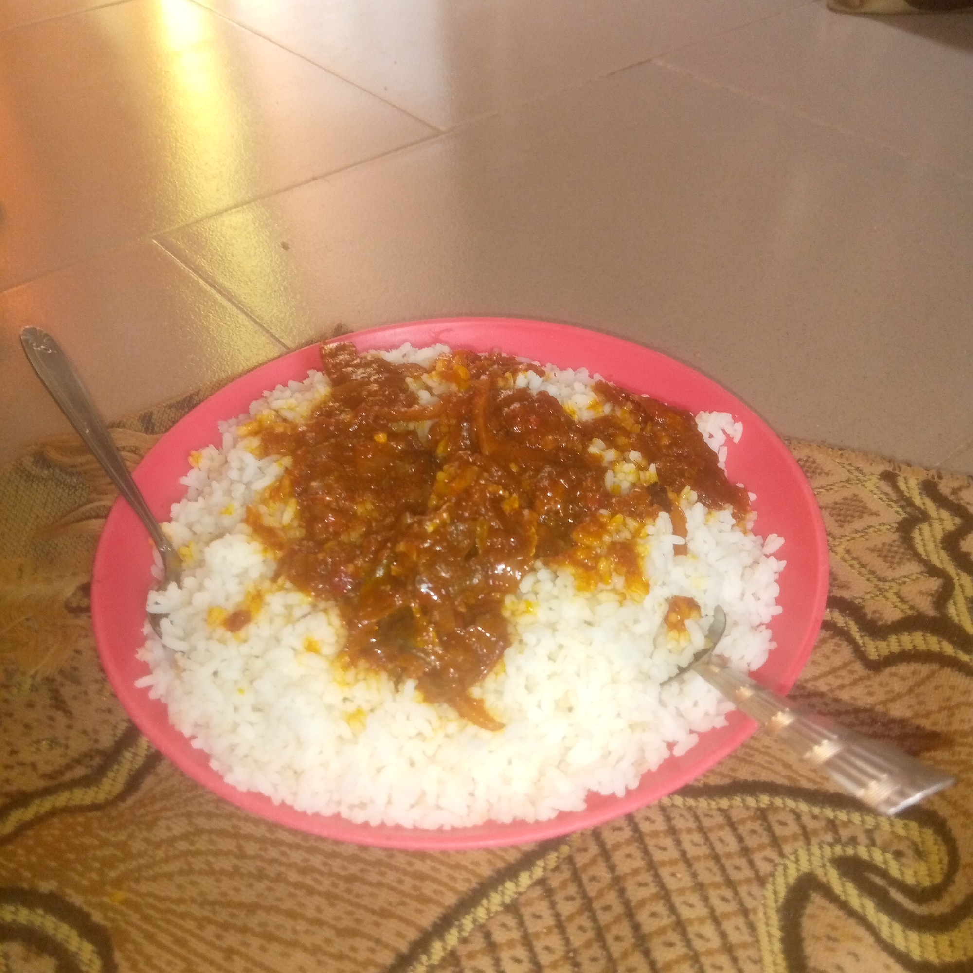 A close-up, slightly blurry, overhead shot shows a pink plastic plate filled with white rice and topped with a reddish-brown stew or sauce. The plate is resting on a brown and yellow patterned fabric, which appears to be a mat or tablecloth. A silver fork is positioned on the right side of the plate, with its tines resting on the rice and stew. A silver spoon is visible on the left side of the plate, partially obscured by the rice. The background consists of a beige tiled floor with a bright light reflection on the upper left portion of the image. There are no people or animals visible in the frame.