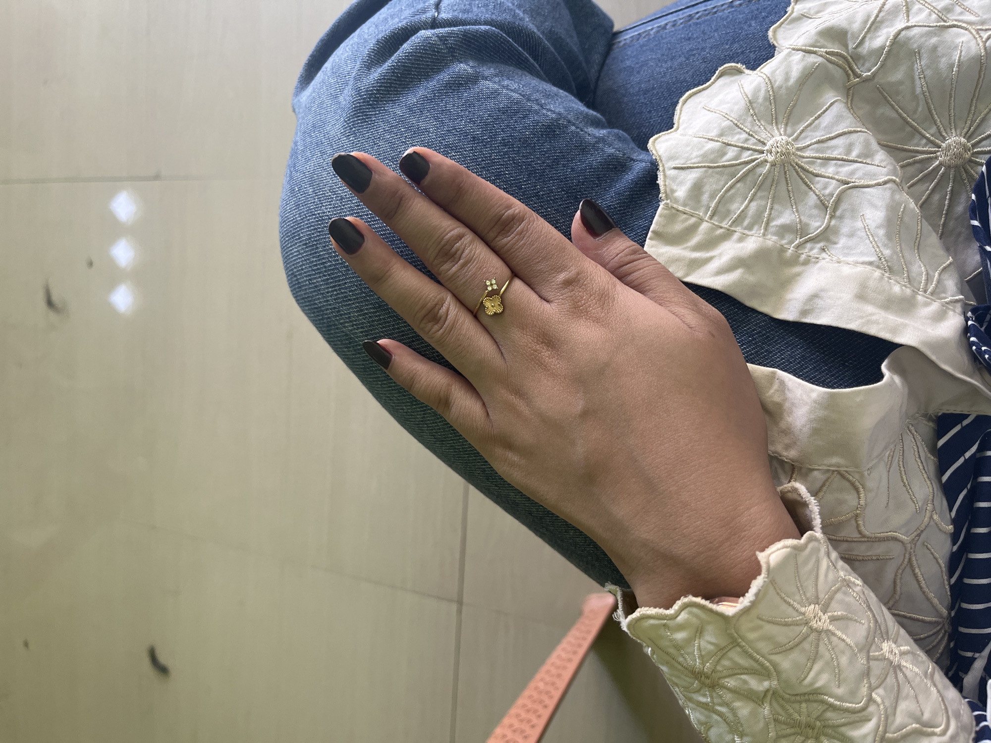 A close-up, overhead shot shows a person's left hand resting on their denim-clad legs. The hand has dark-colored nail polish and is adorned with a delicate gold ring shaped like a flower with small diamond accents. The person is wearing a light-colored blouse with intricate embroidery around the collar and sleeves, and a blue and white striped garment is visible underneath. The background appears to be a light-colored, tiled floor. The scene is likely indoors, given the lighting and the floor surface, and it is not possible to determine the specific location within Bumi Mulyo, Indonesia from the visual information provided.