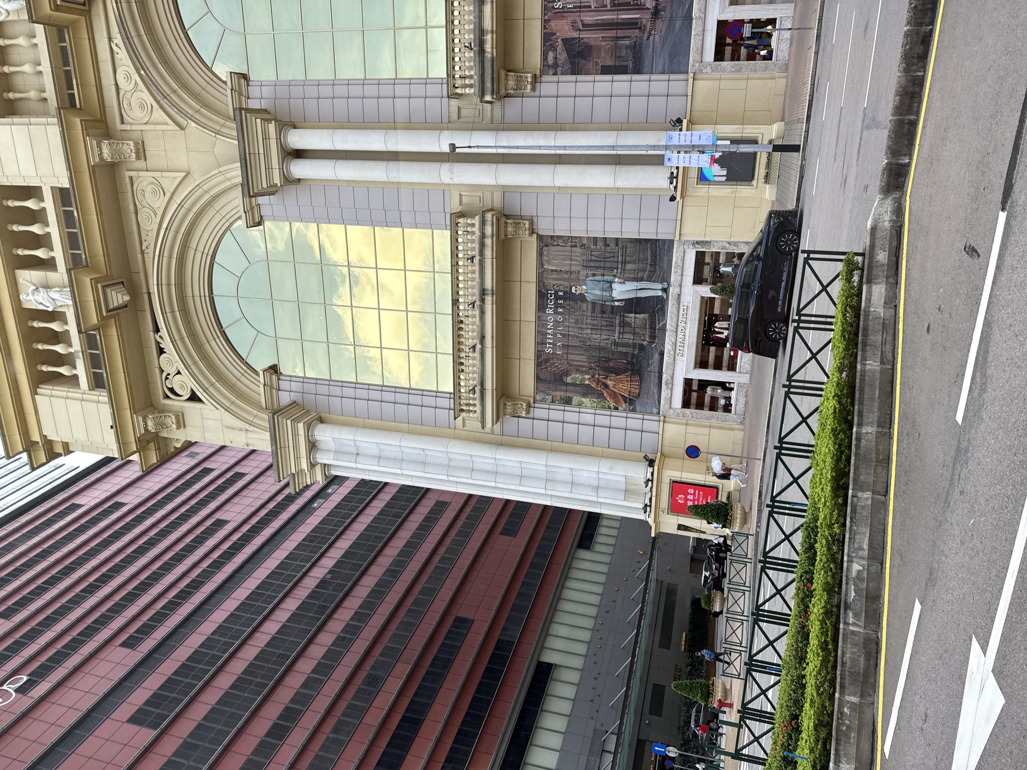 The image shows a street scene in Macau, China, with a large, ornate building on the right and a tall, modern red building on the left. The ornate building has a cream-colored facade with large arched windows reflecting the sky. Below the windows are large advertisements, one featuring a man in a suit. White columns support the upper levels of the building. A black minivan is parked on the street in front of the ornate building, and several people are walking on the sidewalk. A green metal fence separates the sidewalk from a planted area with bushes and small red flowers. The road in the foreground has white lane markings and a yellow line. The red building on the left has a repetitive pattern of horizontal red panels and dark, recessed sections.