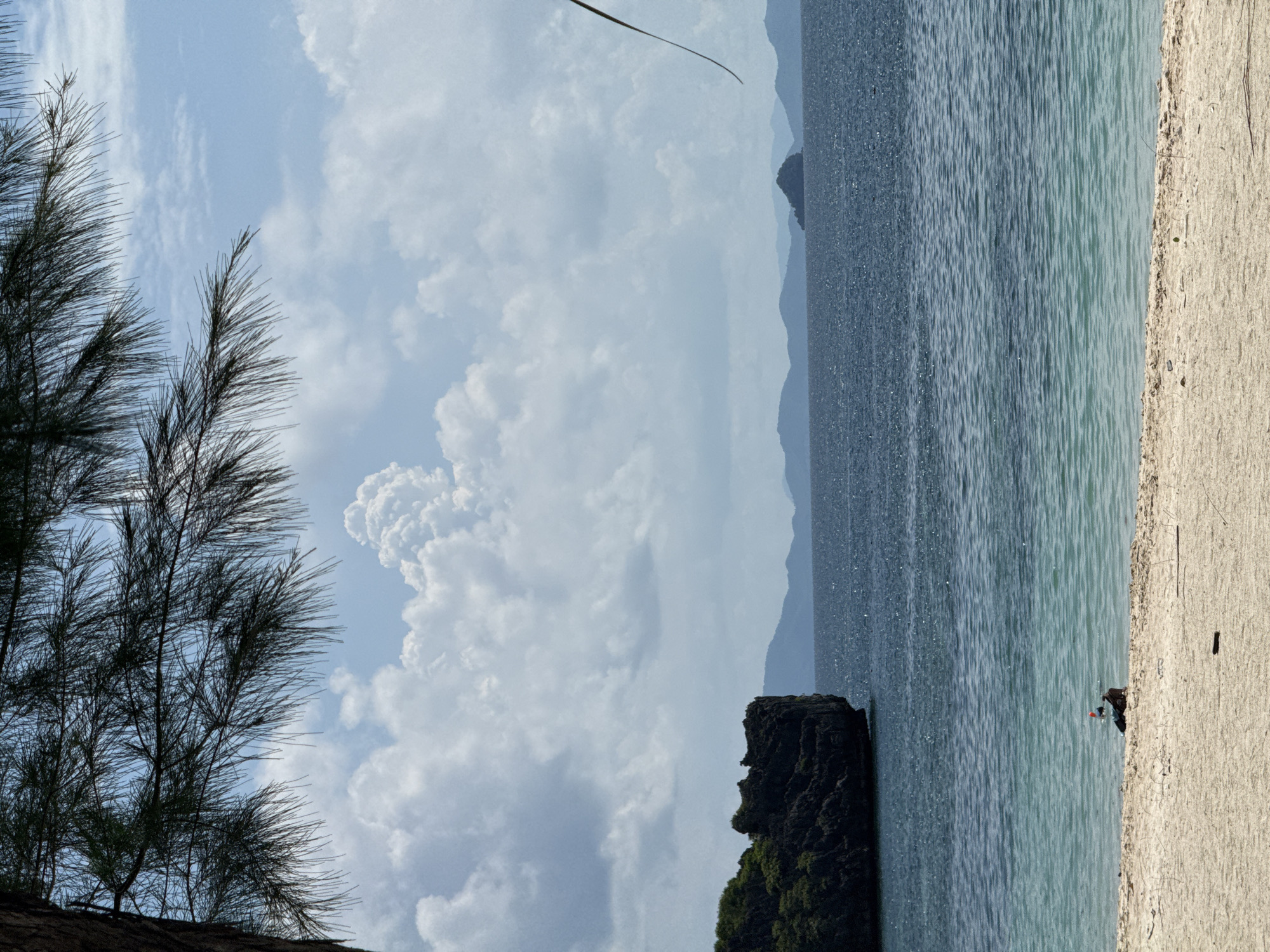 This image depicts a tranquil beach scene in Ban Ao Nang, Thailand. The foreground features a sandy beach with scattered debris, leading to clear turquoise water that transitions into deeper blue. In the shallow water, a single individual is visible, partially submerged and appearing to snorkel or prepare for snorkeling, with snorkeling gear on their head.
The middle ground is dominated by the calm ocean, with a prominent, dark, rocky island formation on the left. In the distance, a chain of hazy, layered mountains forms the horizon, with a small, conical island visible further out at sea on the right.
The sky above is partly cloudy, with large, fluffy white cumulus clouds interspersed with patches of blue sky. The upper portion of the image is framed by the dark, needle-like foliage of a tree branch extending from the left side. The overall impression is one of serene coastal beauty and leisure.