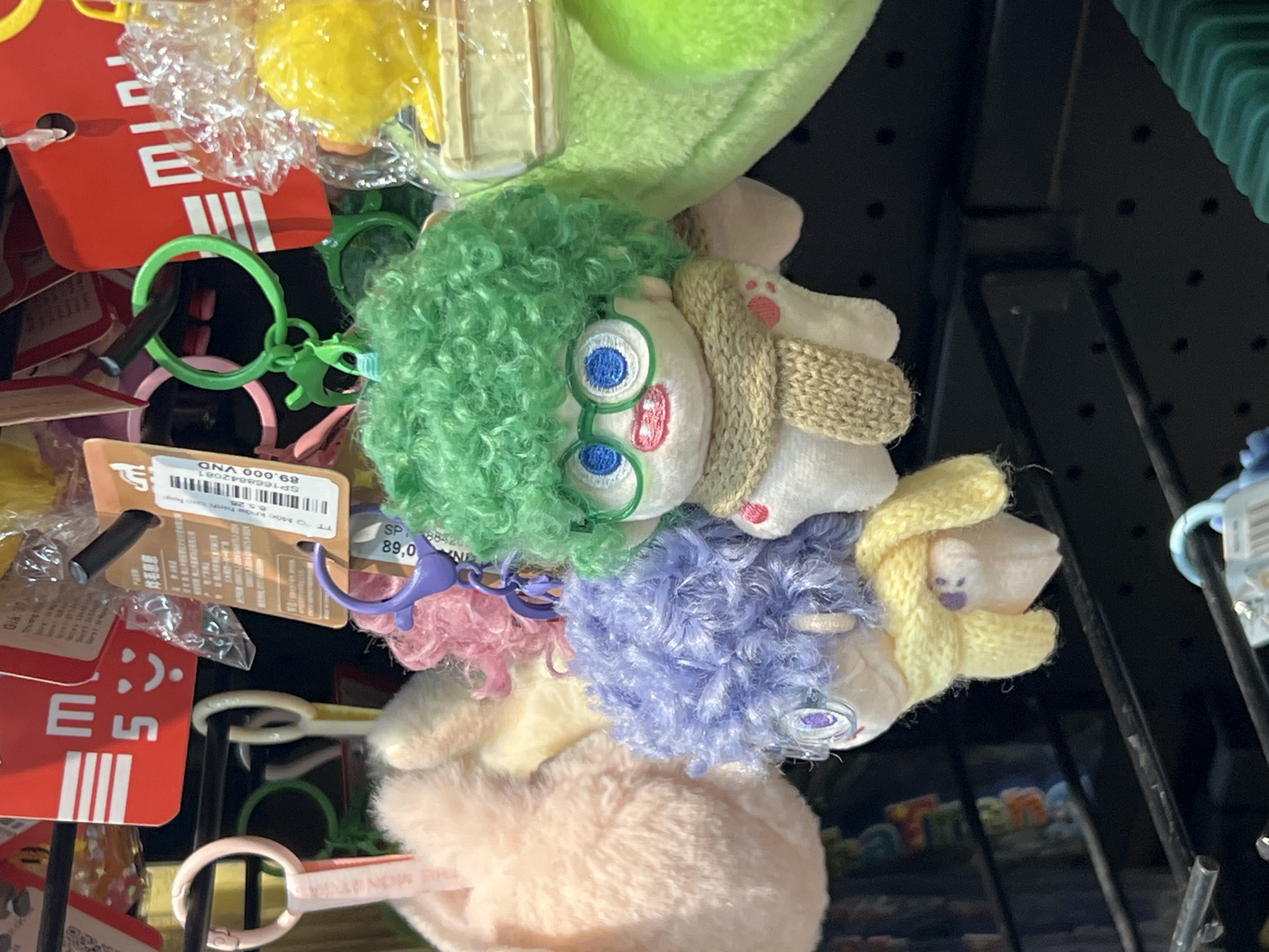 A close-up, top-down shot reveals a vibrant collection of keychains and plush toys displayed for sale, likely in a retail store in Bac Nha Trang Ward, Vietnam. The items are hanging on black metal hooks attached to a black pegboard or grid display rack, partially visible in the background.
The central subject is a whimsical plush keychain featuring a character with unruly, bright green curly hair and a light-colored face. This character wears blue-rimmed glasses, which give the eyes a prominent, almost expressive look, and a light brown knitted scarf around its neck. Its overall appearance suggests a cartoon or anime-inspired design, reminiscent of popular animated characters.
Surrounding this central plush are other keychains of various designs. To the right, another plush keychain is visible, featuring fluffy purple hair and a small knitted yellow appendage. Below it, a lighter pink, very fluffy plush keychain is seen. Other keychains include a bright green plastic keyring with a small animal charm, and a yellow plastic keyring. Red product tags with white lettering are interspersed among the items, along with transparent plastic packaging.
The items appear to be static, displayed for purchase. Notable details include the vibrant colors of the plush hair (green, purple, pink) and the contrasting textures of the fluffy plush and knitted elements. Several tags are visible with text. The most prominent is a white tag displaying "89.000 VND", which is a price in Vietnamese Dong, confirming the location context. Other partial texts include "SP1668842081" (likely a product code), "Gía 30" (Price 30), "THE MONST" (possibly part of "The Monsters"), "ONA", "ES", and "6.5.20" (possibly a date). The scene evokes a cheerful, commercial atmosphere typical of gift shops or souvenir stalls.