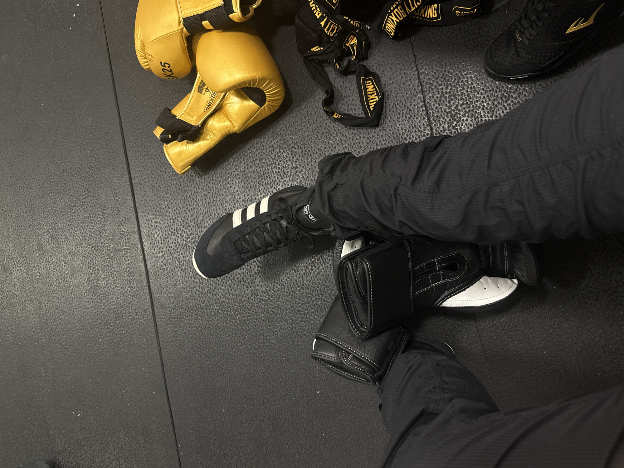 The image captures an overhead view of a textured, black rubber gym floor, typical of a boxing or martial arts training facility. Several pieces of boxing equipment are arranged on the floor, alongside the legs and footwear of at least two individuals.
In the upper left quadrant, a pair of shiny golden boxing gloves is visible, one showing the partially legible text "KING" and "K25", along with "BOXING". Adjacent to these gloves, a black hand wrap with distinctive gold lettering is loosely placed, displaying "CITY BOXING" and "CAMP" in a repeated pattern.
Towards the center and lower right, a person's lower leg and foot are seen, clad in dark, possibly athletic, trousers and a black Adidas sneaker with prominent white stripes and the "adidas" logo on the tongue. Below this, a pair of black and white boxing focus mitts or pads rest on the floor. Further to the right in the upper section of the image, another person's foot, wearing a black athletic shoe with a gold logo (resembling Everlast's "E" or similar) and partial text, is visible.
The scene suggests a moment before, during, or after a boxing or combat sport training session. The presence of multiple types of boxing gear and two sets of legs indicates at least two participants. No specific time of day or weather cues are discernible due to the indoor setting. The visible text confirms brands like "CITY BOXING," "KING," "adidas," and potentially "EVERLAST."