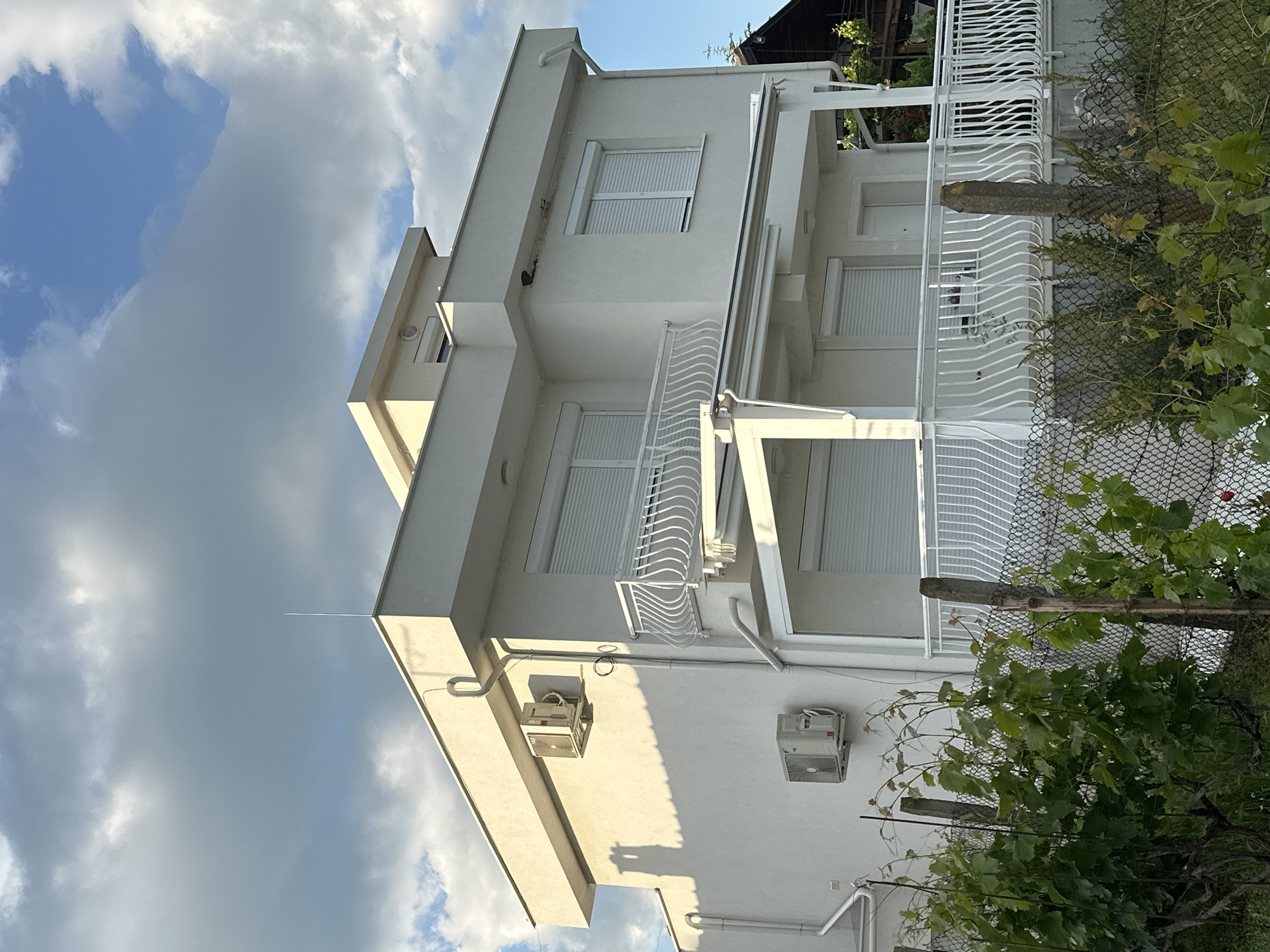 The image shows a white two-story building, likely a house, under a partly cloudy sky. The building has rectangular windows with white blinds and white balconies with wavy metal railings. There are two air conditioning units visible on the side of the building. A black wire fence partially obscures the right side of the building, with some greenery growing along it. The location is not identified in the image.