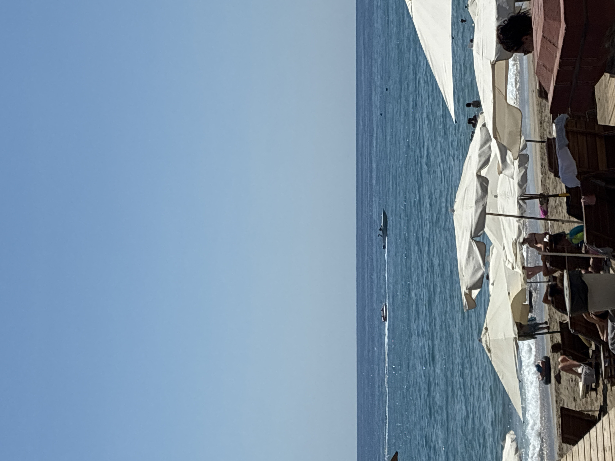 The image depicts a sunny day at a beach in Alanya, Turkey. The foreground shows a sandy beach with several white beach umbrellas and lounge chairs. Some people are visible relaxing on the chairs and walking on the beach. In the middle ground, the sea is calm with gentle waves lapping the shore. Two speedboats are seen in the distance on the water. The sky is clear blue, indicating fair weather. The scene suggests a typical summer day at a popular tourist destination.