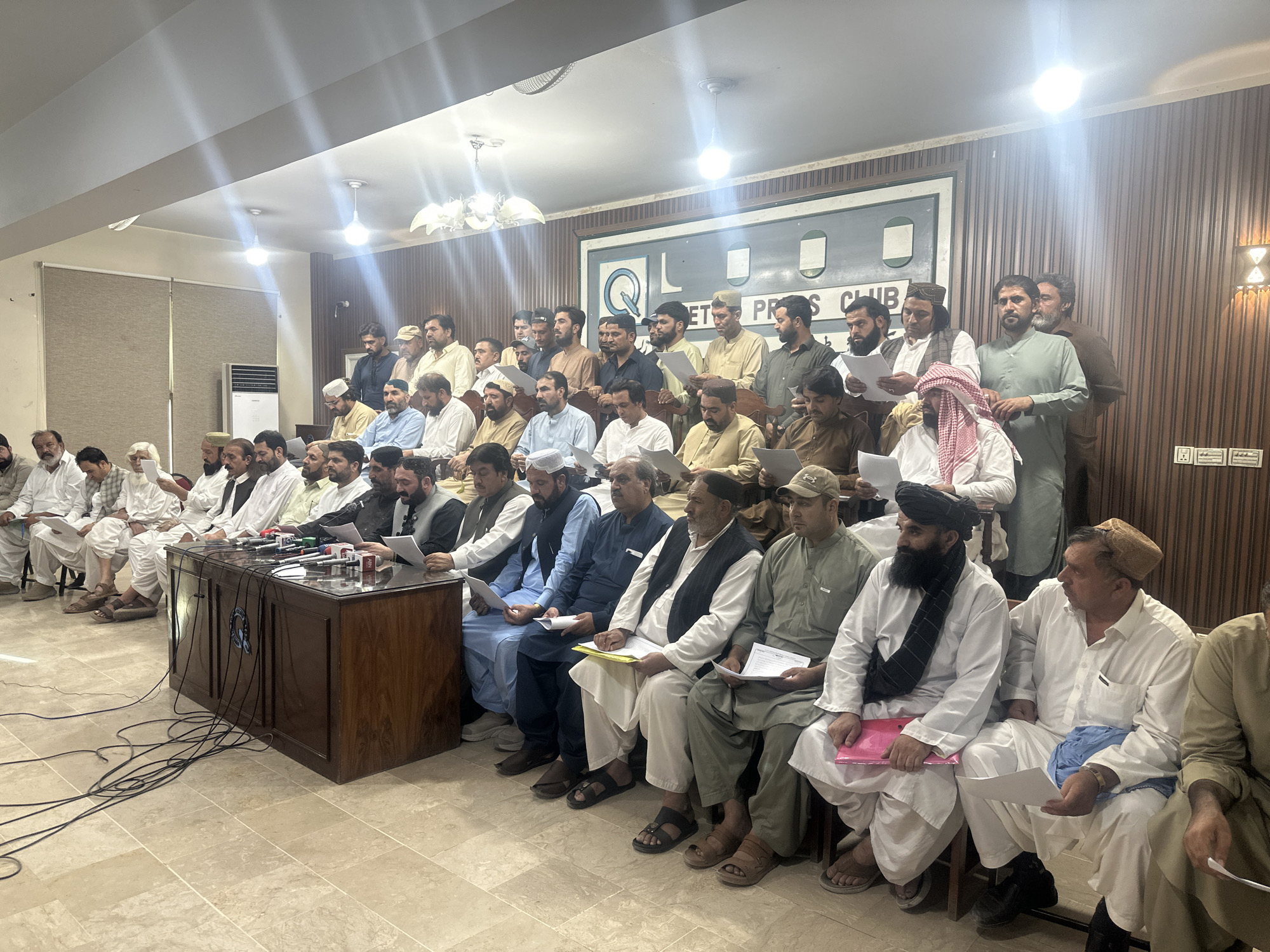 A group of approximately thirty-five men are seated in rows in what appears to be a press conference or meeting room. The men are dressed in traditional South Asian attire, primarily shalwar kameez, with some wearing vests or caps. Many are holding papers, and some are looking towards a wooden podium in the foreground which is equipped with multiple microphones. Behind the seated individuals, a large banner is visible with text that includes "PRESS CLUB". The room has a tiled floor and paneled walls, with lighting fixtures on the ceiling. An air conditioning unit is visible on the left side of the image. The overall setting suggests a formal gathering in Quetta City Tehsil, Pakistan.