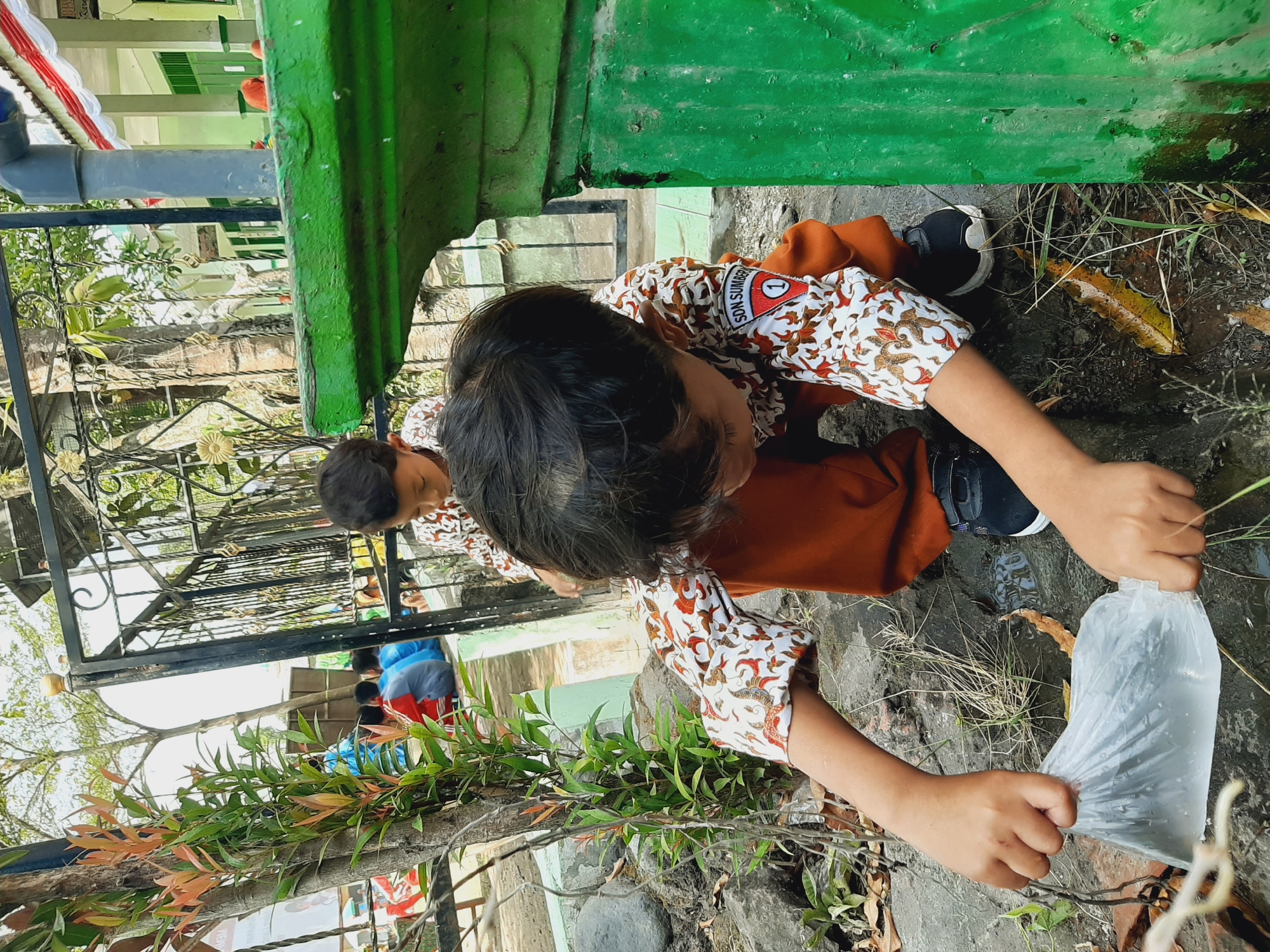 In Sumberjo, Indonesia, two young boys in school uniforms are engaged in an activity outdoors. The boy in the foreground, wearing a white shirt with an orange and brown batik pattern and orange pants, is crouching down. He is holding a clear plastic bag containing a dark object, possibly a fish or a small animal. His hands are gripping the top of the bag. Beside him, another boy, also in a similar uniform, is visible in the background, also crouched. They are in an outdoor area with a stone pathway, some vegetation, and a green structure that appears to be part of a building or a fence. A metal gate is visible behind the boys. The environment suggests a schoolyard or a public space.