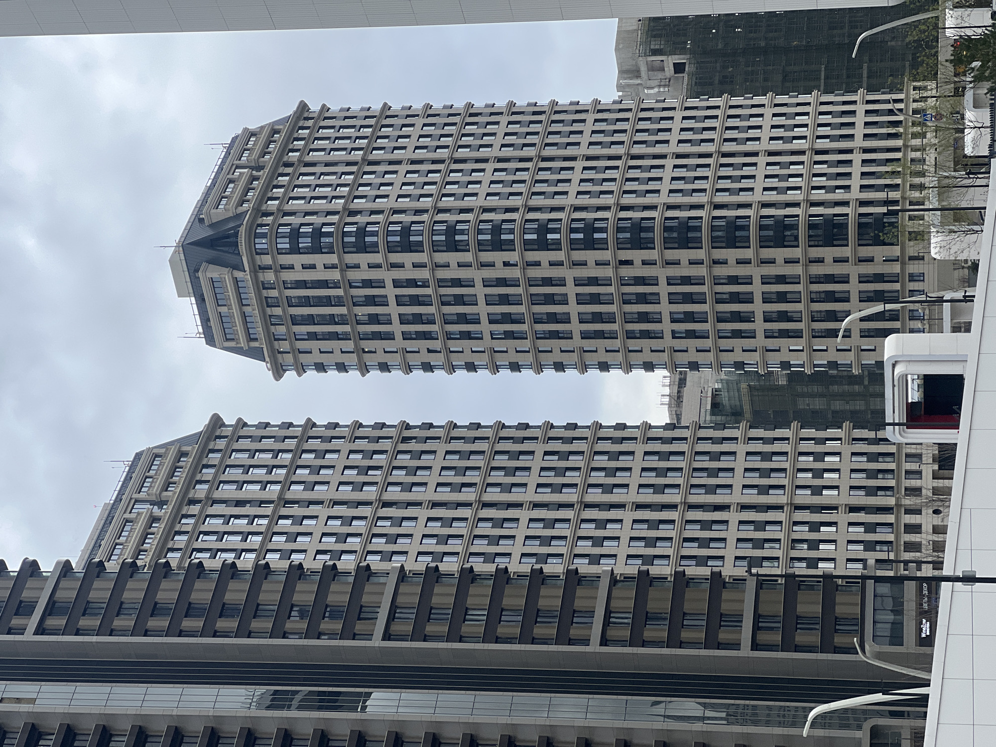 The image shows two tall buildings, likely office or apartment complexes, with a distinctive architectural style featuring regularly spaced rows of windows and prominent horizontal lines. The buildings are constructed of light-colored stone or concrete with dark window frames. The sky is overcast. Other buildings are visible in the background, including one under construction and another partially obscured by the buildings in the foreground. Street lights and a portion of a building with signage are also visible. The image is not taken in Khimki, Russia.