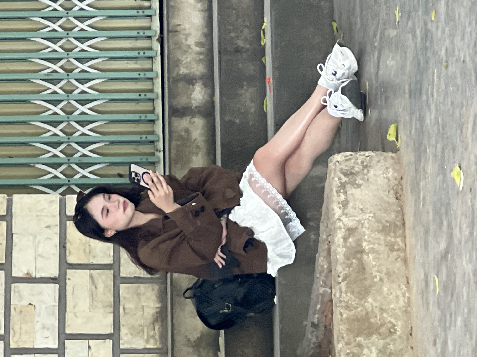 A young Asian woman with long dark hair and light makeup sits on a concrete staircase, looking at her smartphone. She is wearing a brown jacket over a white lace-trimmed dress and white sneakers. To her left is a black backpack. The staircase appears to be located outdoors, possibly in a public area of Da Nang, Vietnam, as indicated by the barred green gate behind her and the brick wall to her left. Scattered yellow leaves are visible on the stairs and the ground.