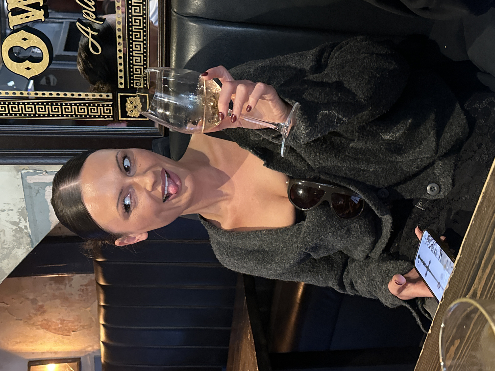 A woman is seated indoors, likely in a pub, bar, or restaurant in London, United Kingdom, at a dark wooden table in a leather booth. She is looking towards the camera with a playful expression, her tongue slightly out, and a smile on her face. Her dark hair is pulled back, creating a sleek appearance.
She wears a dark grey, textured cardigan, with black sunglasses casually tucked into its neckline. Her left hand holds a clear wine glass containing a light-colored liquid, and her fingernails are painted a dark red or burgundy. In her right hand, she holds a smartphone, which displays an image of what appears to be a sword.
The background features dark leather booth seating. Behind her to the left is a large, ornate mirror with prominent gold detailing. Visible elements on the mirror include the partial text "A pla", a large gold "8" or similar symbol, a Greek key pattern border, and a small golden owl emblem. The surrounding walls exhibit a distressed or textured finish, with varying tones of light grey/white and reddish-brown. The lighting in the scene is dim, suggesting an evening setting. The overall atmosphere is casual and social.