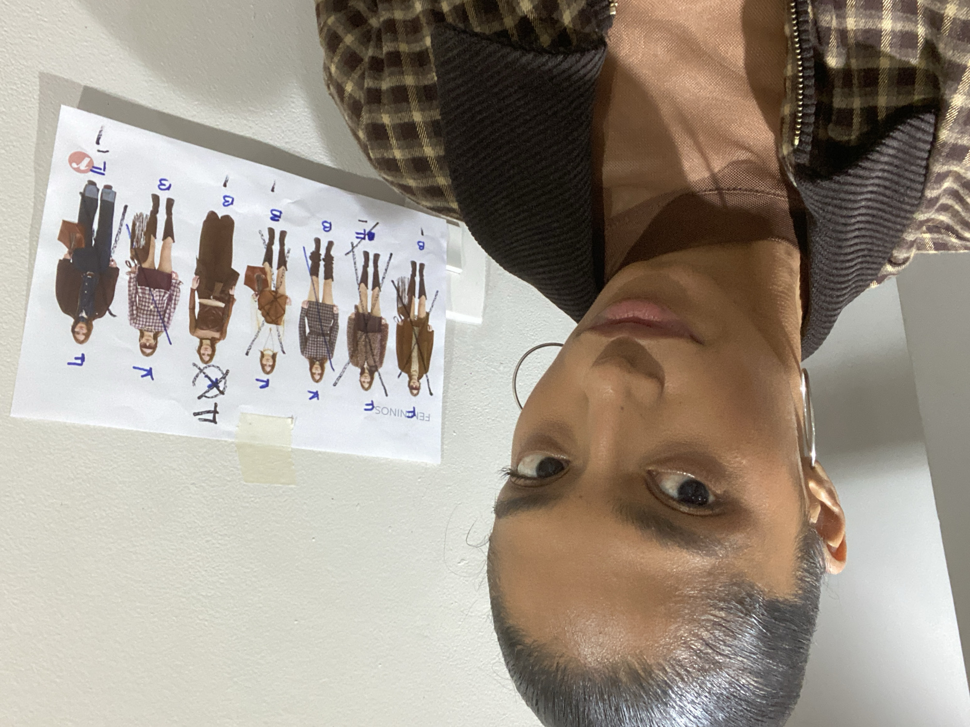 A selfie-style, slightly distorted image shows a person with dark hair pulled back, wearing large hoop earrings and a brown plaid jacket over a sheer top. Their face is angled towards the camera, displaying one eye prominently. Beside them is a sheet of paper taped to a white wall. The paper features cartoon-like depictions of people dressed in various outfits and marked with handwritten annotations in blue ink. The words "FEININOS F" are also printed on the paper. No specific details indicate that this image was taken in São Paulo, Brazil.