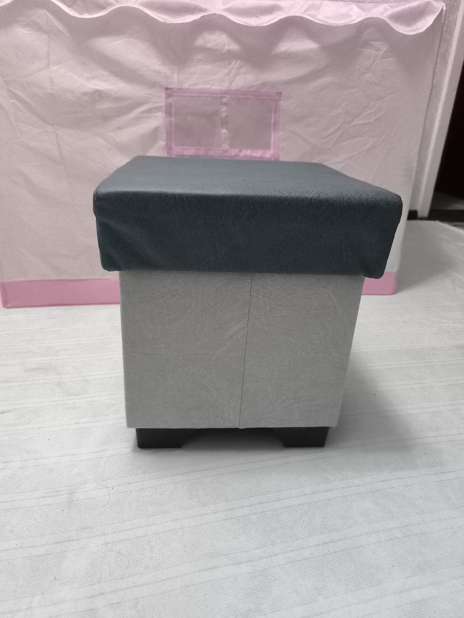 This media file showcases a close-up, eye-level view of a square ottoman with a dark gray cushioned top and a light gray textured base. The ottoman has a sturdy appearance, supported by four dark, blocky feet.
In the background, a large, soft pink tent-like structure is visible, featuring a scalloped edge along its top and a transparent rectangular window with a pink trim. The tent is made of a lightweight, possibly fabric material, with visible creases and folds suggesting its pliable nature. A thin, horizontal pink band is present along the lower edge of the tent.
The scene is set on a white, textured surface, possibly a rug or fabric floor covering, with subtle linear patterns. The lighting is even, suggesting an indoor setting with diffused light, and there are no strong shadows. The overall impression is one of a clean, domestic environment. No people are visible in the frame. The image does not contain any visible text. The context provided indicates the location as Kokand, Uzbekistan.