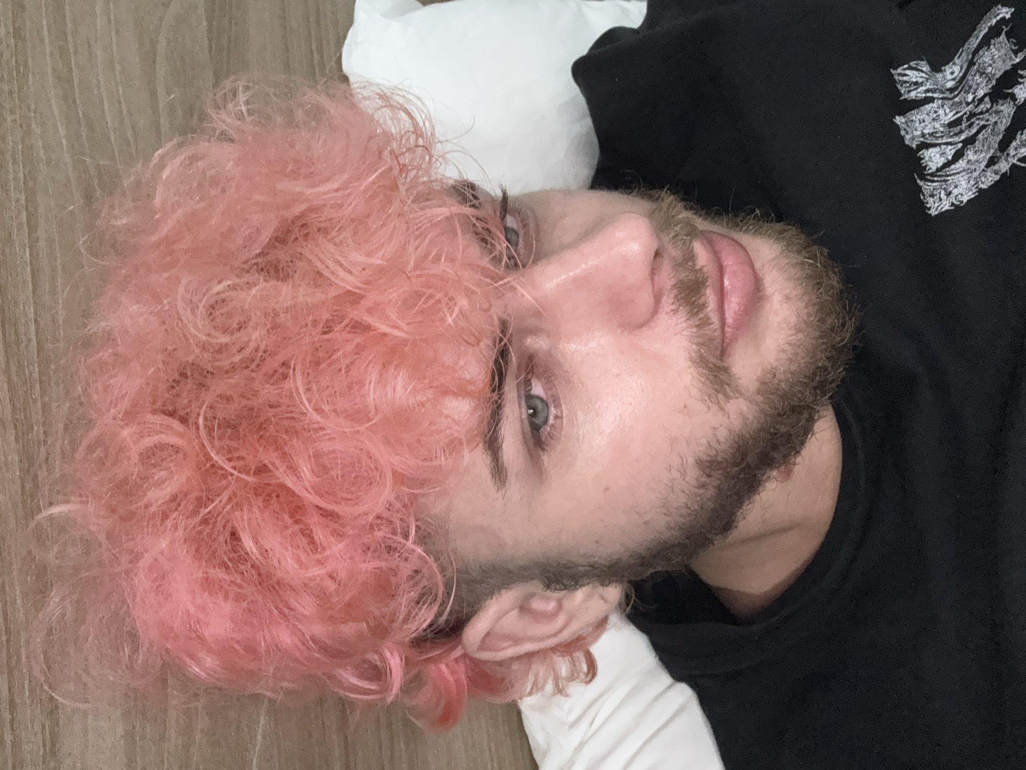 This media file is a close-up, portrait-style image of a person lying on their back indoors. The subject has distinctive curly, bright pink hair that spills onto a light wooden floor. They possess light-colored eyes and a light brown beard or stubble. The person is looking directly at the camera with a neutral and calm expression.
They are wearing a black t-shirt or top, with a white, abstract design visible on the upper right portion of the garment, which appears to be a stylized pattern rather than legible text. A white pillow or fabric is visible beneath the person's head, contrasting with the dark shirt and pink hair. The background consists of a light-toned wooden floor or paneling, indicating an indoor setting. The image is oriented with the subject's head facing the right side of the frame. No specific activity is taking place beyond posing for the photograph. The location context is Đà Nẵng, Vietnam.