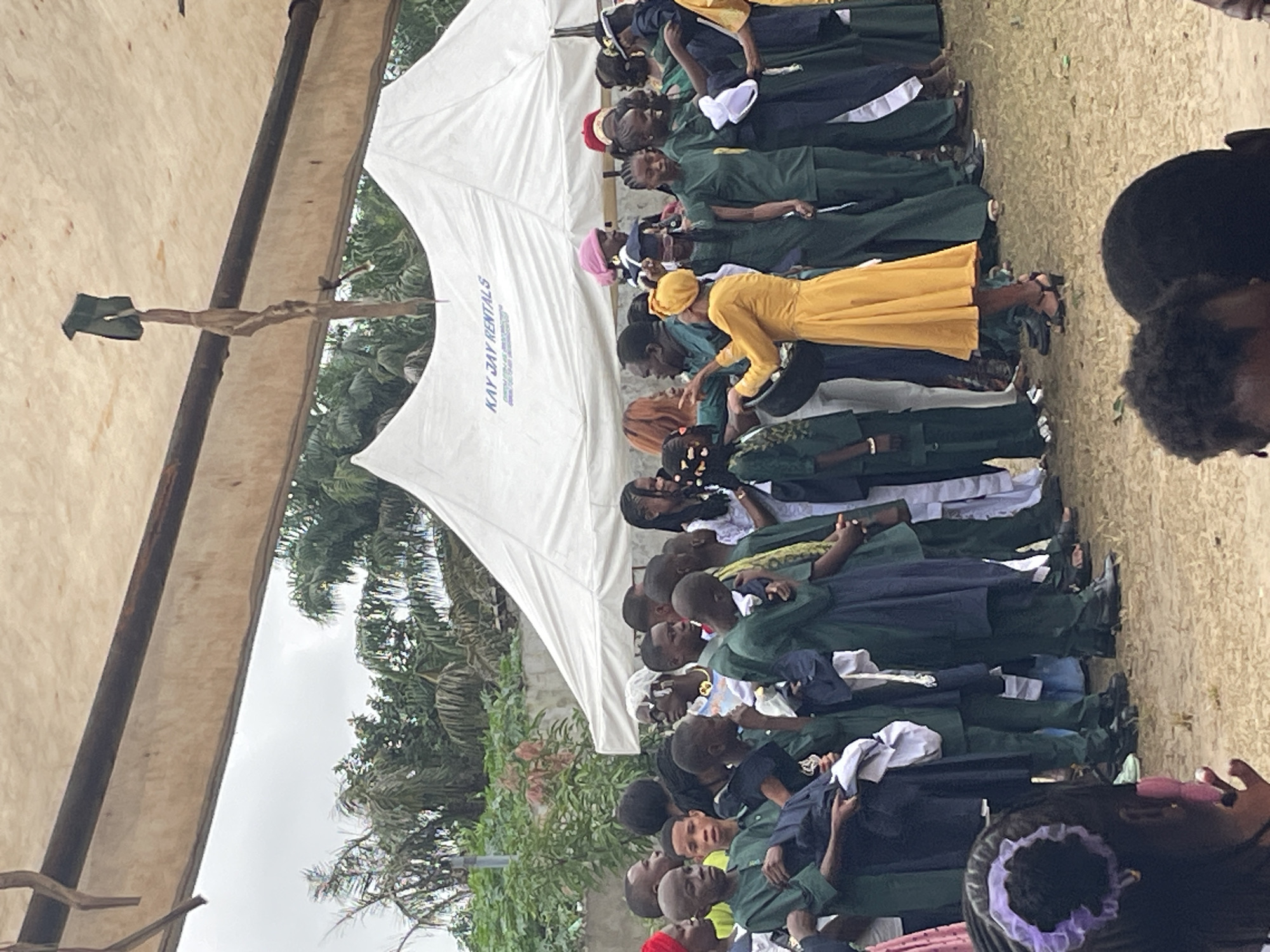 This image depicts an outdoor gathering in Badagry, Nigeria, likely a school or community event, under a large white tent that bears the inscription "KAY JAY RENTALS." A diverse group of people, including many children and young adults, are assembled. Many of the younger attendees are dressed in matching dark green uniforms, suggesting a formal occasion or a specific organization. Some individuals are wearing brightly colored clothing, such as a woman in a vibrant yellow dress who appears to be interacting with a group. The background reveals lush greenery and palm trees, characteristic of the Nigerian landscape. The overall atmosphere seems to be one of celebration or a structured event, with people standing in rows and engaging with each other. A wooden structure with a torn cloth hanging from it is visible suspended from the tent's frame, adding an element of rustic decoration or perhaps functional use. The ground is covered in sand and dirt, indicating an outdoor setting.