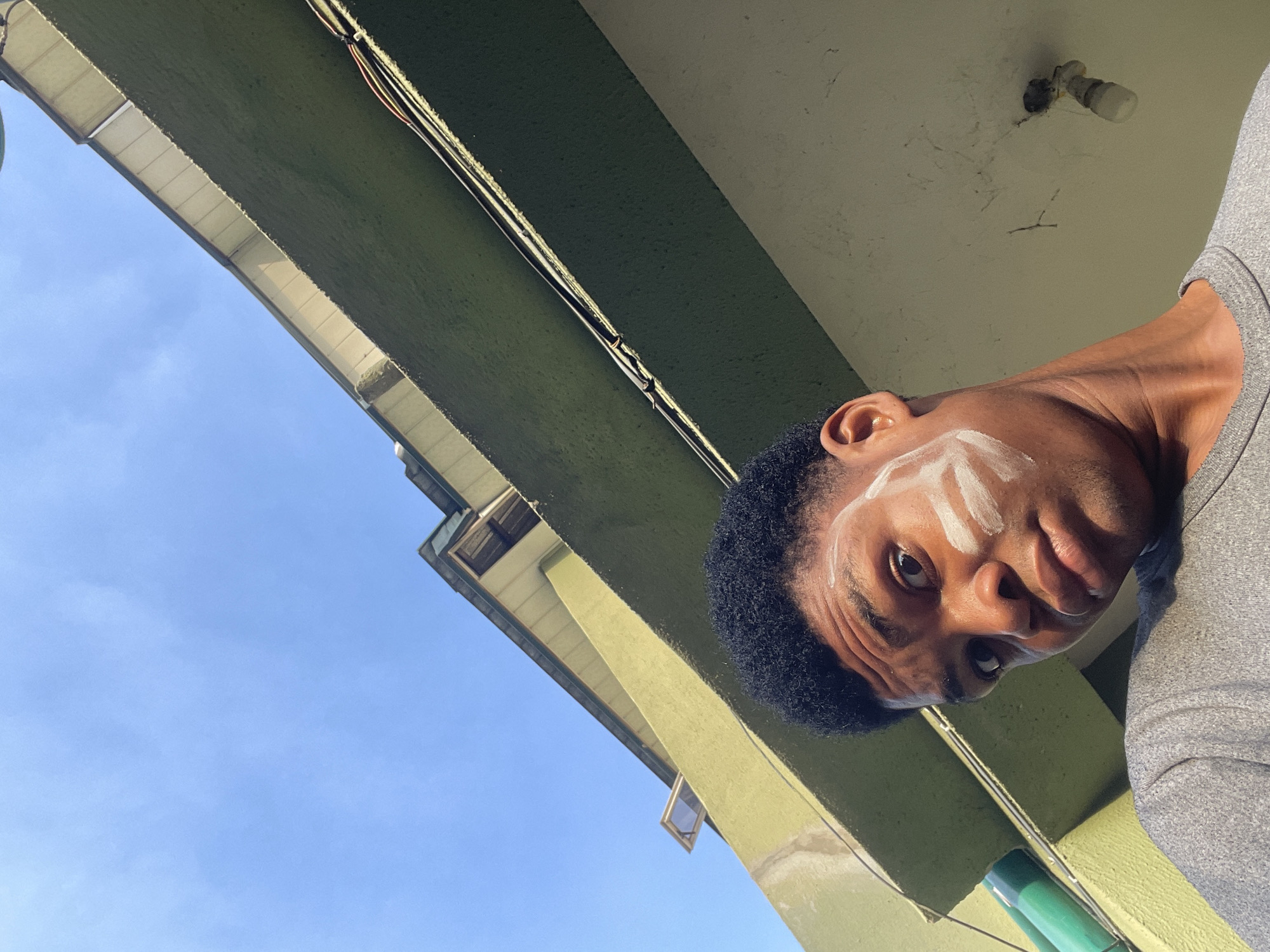 A low-angle shot depicts a man in Ado, Nigeria, with white markings on his face, possibly tribal. The view is from below, looking up at him against the backdrop of a building with green overhangs and a patch of blue sky. The building's architecture, including the windows and construction materials, is visible. Electrical wires run along the building. The man is wearing a gray shirt and has short, dark hair.