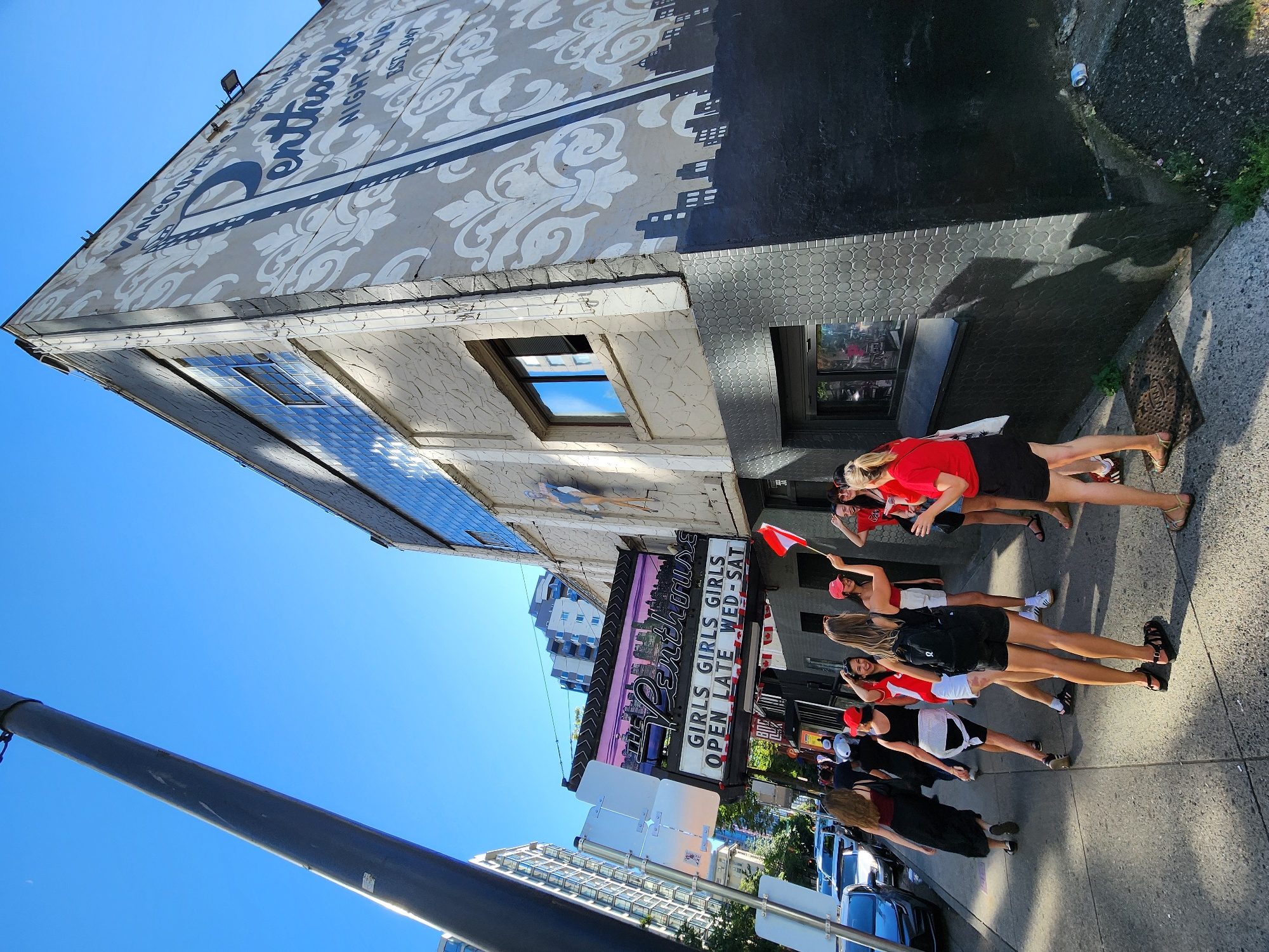 The image shows a group of young women gathered on a sidewalk outside a building. The building has a large sign that reads "Penthouse Girls Girls Girls Open Late Wed-Sat." The building's facade is decorated with a mural of a cityscape and ornate patterns. The women are dressed in casual clothing, some wearing red tops and others in black outfits. One woman is holding a small Canadian flag. Several cars are parked along the street. The scene takes place on a sunny day with a clear blue sky. The environment appears to be an urban setting, possibly a commercial or entertainment district.