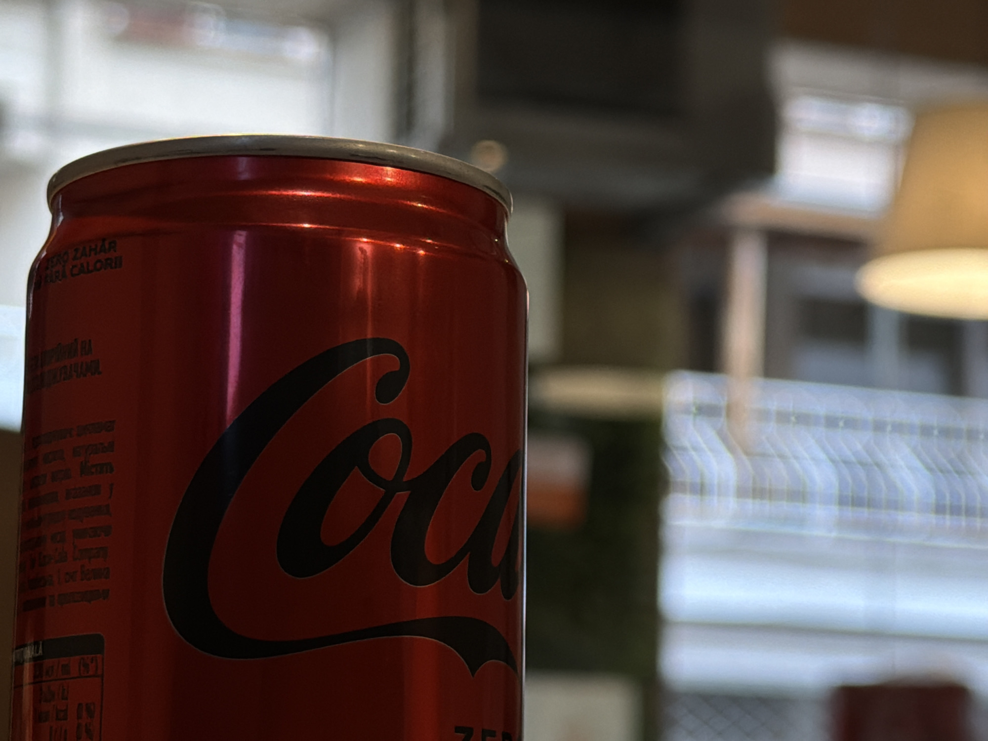 A red Coca-Cola Zero aluminum beverage can is positioned in the left foreground, in sharp focus. The can features the brand's iconic black script, with the word "Coca-Cola" prominently displayed on its lower body. Cyrillic text, including "ZERO ZAHÁR" (zero sugar), is visible on the left side of the can's surface. The top portion of the can, including its silver rim, is partially cropped from the image. The background is significantly blurred, indicating an indoor setting. To the right, a warm-toned, blurred lampshade or light fixture is visible, suggesting ambient lighting. Further right, a vertical pattern of blurred white lines, possibly from window blinds or a paneled structure, is discernible. The overall environment appears to be a commercial establishment, such as a cafe or restaurant, located in Sofia, Bulgaria, as indicated by the Bulgarian text on the can. No individuals, actions, or interactions are present in the scene.