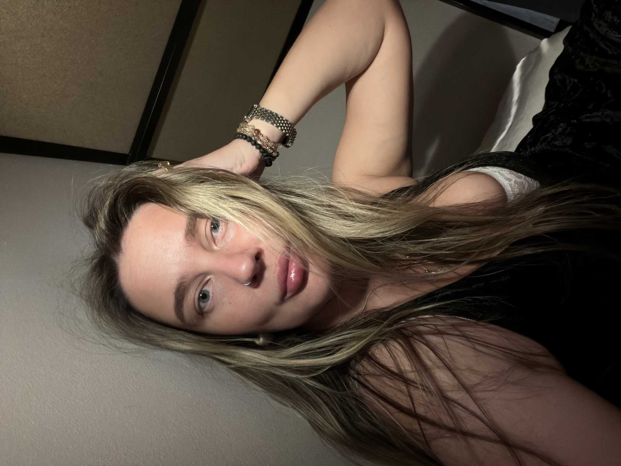 A woman with long blonde hair is lying down, her head resting on a gray surface. She is wearing a black top and a white lace bra is visible. Her left arm is raised above her head, adorned with a collection of bracelets. The background is dark with hints of a bed and a frame. The location is not discernible from the image.