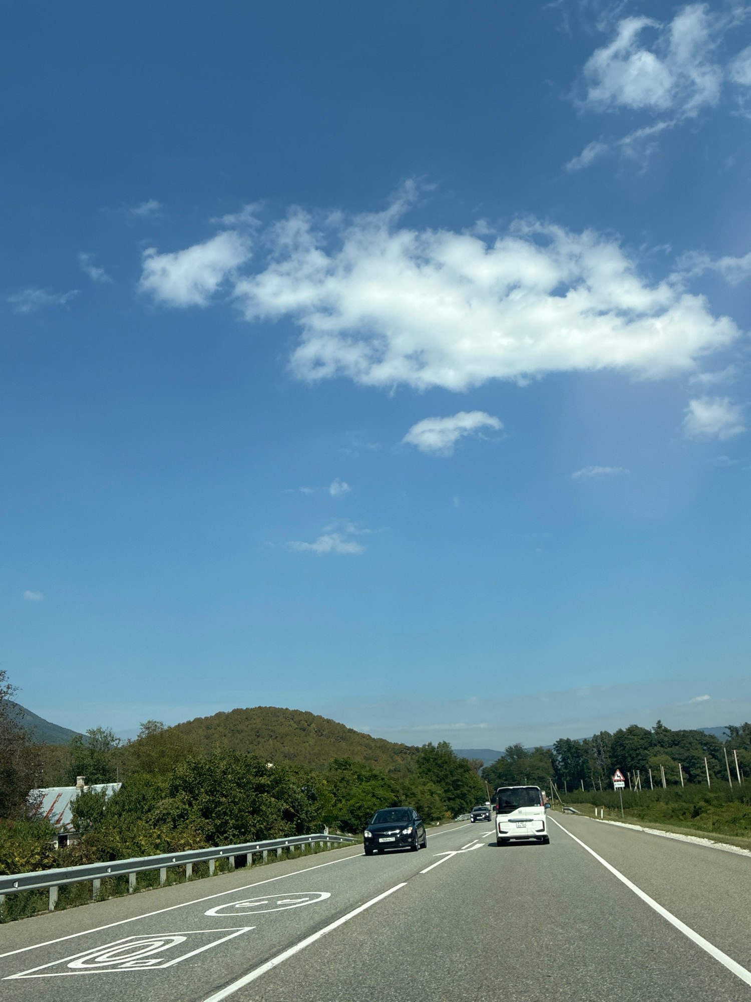The image captures a view of a highway in the city of Shirokaya Pshadskaya Shchel', Russia, on a clear day. The sky is a bright blue with scattered white clouds. The road has multiple lanes and is marked with white lines. Speed limit signs are visible on the road surface, indicating 50 and 20. Several cars are driving on the highway, including a black sedan, a white van, and other vehicles further down the road. On the left side of the highway, there is a metal guardrail, followed by trees, a small building with a gray roof, and a tree-covered hill in the background. The right side of the highway is lined with trees and a signpost. The overall scene depicts a typical day on a Russian highway with cars in motion and the natural landscape surrounding it.