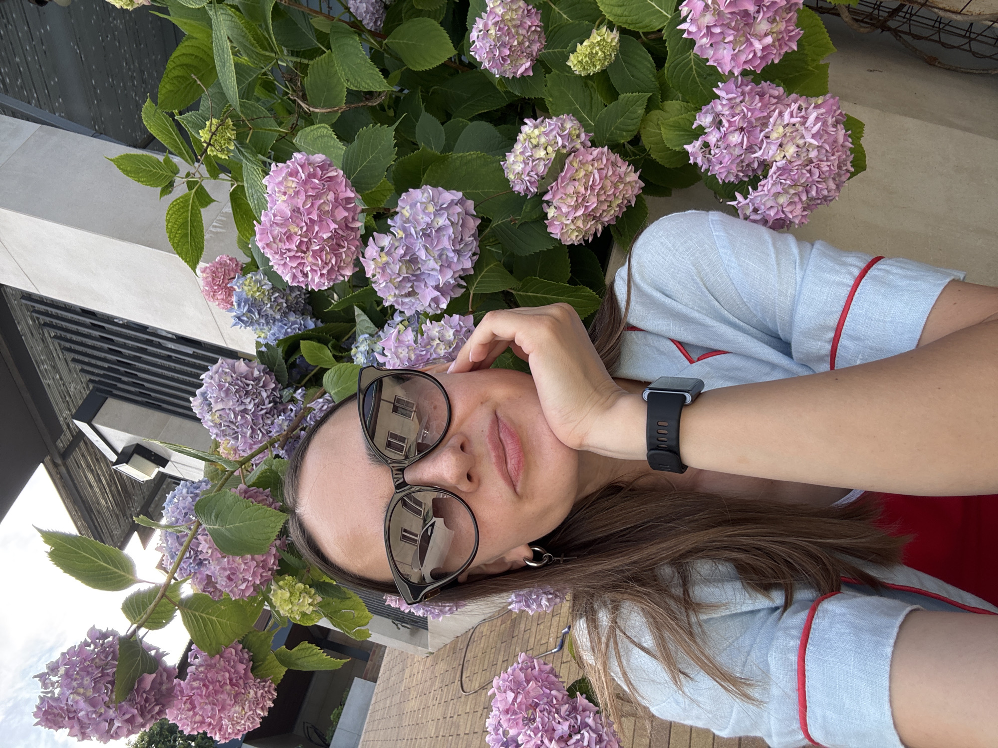 A woman with brown hair, wearing large dark sunglasses, a light blue pajama-style top with red trim, and a red undershirt, is pictured outdoors. She is resting her chin on her hand, with her arm bent. The background features abundant hydrangeas in shades of pink and purple, lush green leaves, and a portion of a building with a modern, angular architectural design. Part of a tiled roof is visible in the upper left corner. The overall scene suggests an outdoor setting, possibly a garden or patio area. The image quality is high, with clear detail.