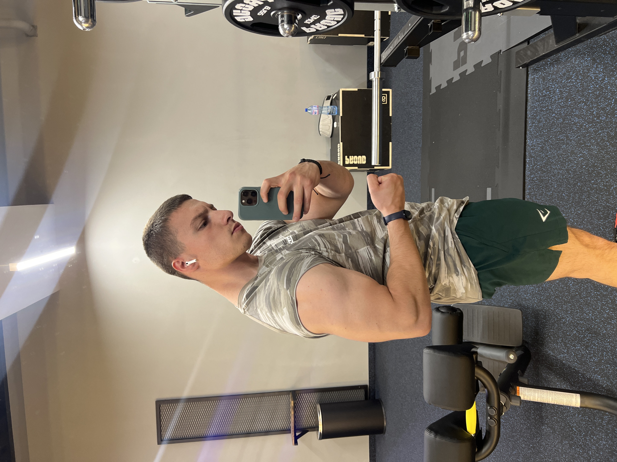 A man is in a gym, holding a cell phone in his left hand, flexing his right bicep. He is wearing an earbud in his left ear, a camouflage t-shirt, green shorts, and a fitness tracker on his left wrist. Gym equipment is visible in the background. The location is not specified, but the city is Burgas, Bulgaria.