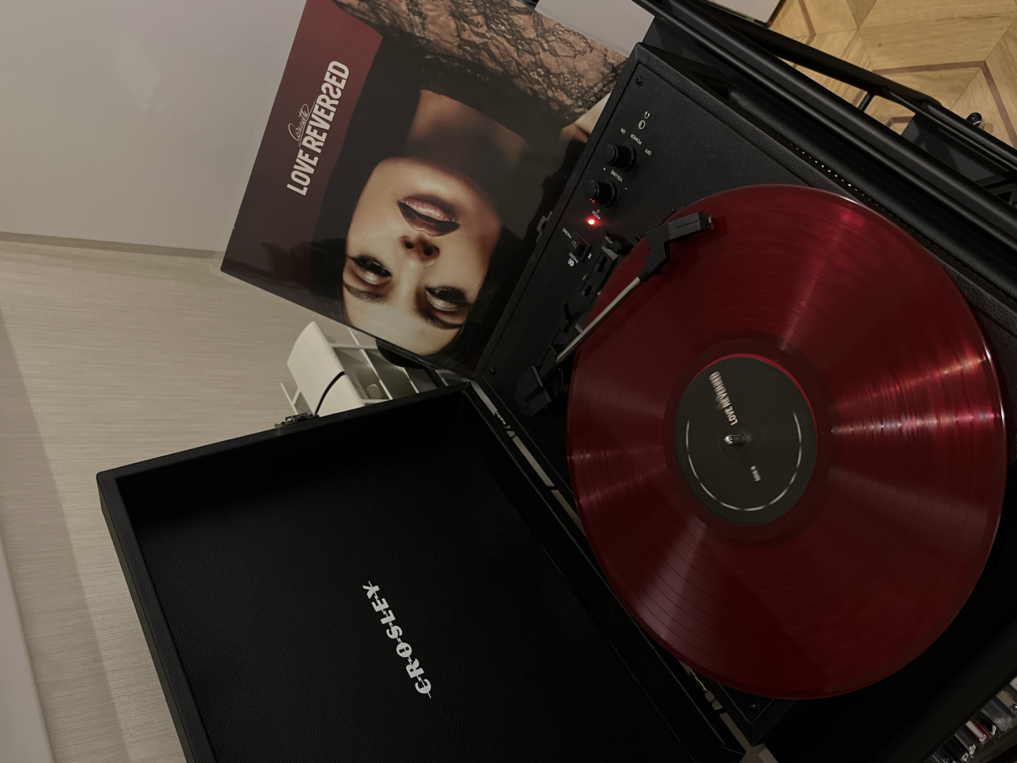 The image shows a Crosley brand turntable playing a red vinyl record. The album cover, titled "Love Reversed" by "Corrinette," is placed next to the turntable. The turntable is black and has a red indicator light. The vinyl record is red with a black label. The overall environment appears to be a room with parquet flooring, but there is no indication of the location in Yerevan, Armenia.