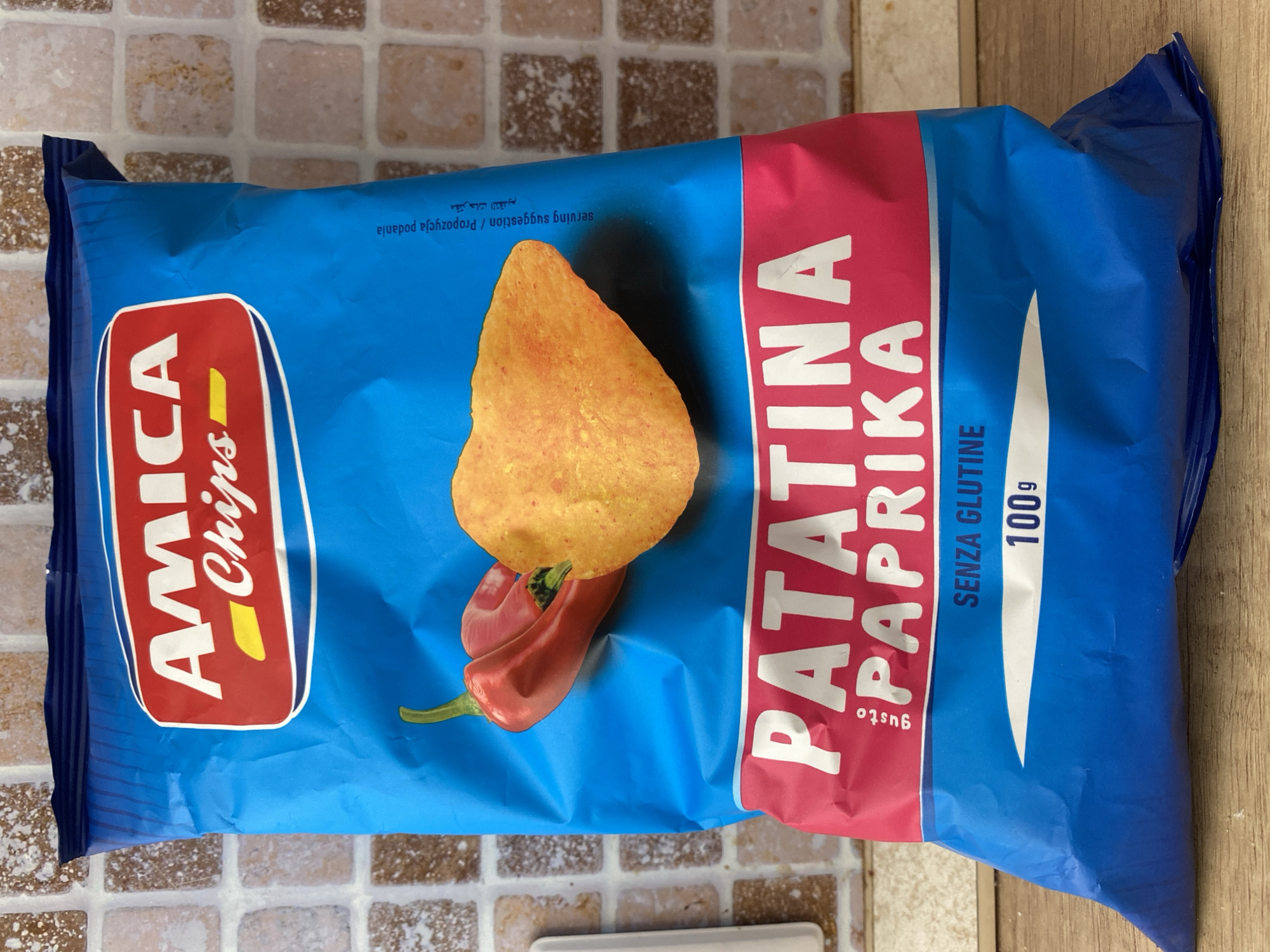 The image shows a bag of "AMICA CHIPS PATATINA PAPRIKA" potato chips. The bag is predominantly blue with a red and white logo at the top that says "AMICA Chips". Below the logo, two red chili peppers and one potato chip are depicted. The bottom portion of the bag has a pink band with "PATATINA PAPRIKA" written in white letters. Below this, "SENZA GLUTINE" (gluten-free) and "100g" are visible. The bag is placed on a wooden surface against a tiled wall. There is no indication of the city of Chișinău, Moldova, in the image.