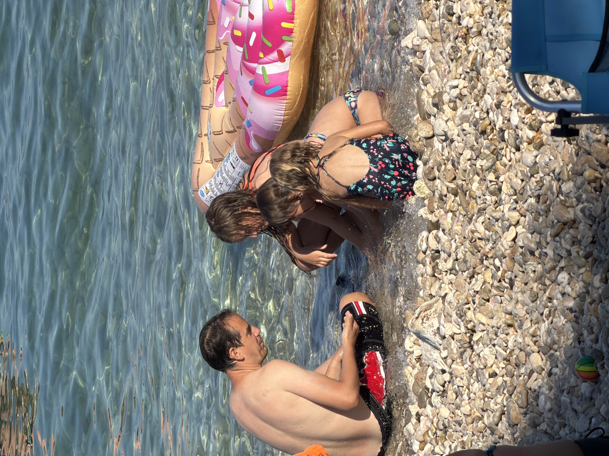 In Općina Baška, Croatia, on a pebble beach, a shirtless man in black swim trunks with red and white trim sits in shallow water. He is looking down at his hands, which are holding a small, yellow object. Near him, three children are also in the water. The two older children, a girl and a boy, are partially obscured, but the girl appears to be wearing a patterned swimsuit. The youngest child, a girl, is facing away from the camera, also in a patterned swimsuit. Behind them, a large, inflatable donut-shaped pool float is partially submerged in the water. The water is clear and shallow, with sunlight reflecting off its surface, and the seabed is covered in smooth pebbles.