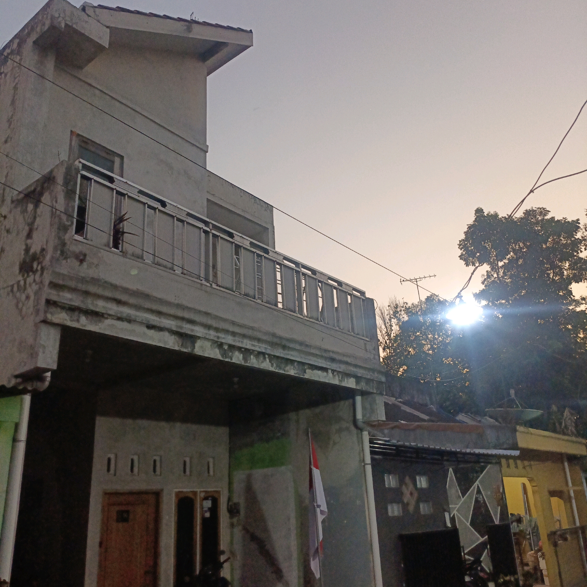 The image depicts a residential building in Salatiga, Indonesia, at dusk. The building is multi-story with a concrete facade, showing signs of wear and tear. A balcony with a metal railing is visible on the upper level, with a plant peeking out from behind the railing. The lower level features a wooden door and what appears to be another entrance or window. A small Indonesian flag is mounted on a pole on the lower right side of the building. In the background, trees are silhouetted against the fading sky, and the sun's glare is visible behind them. Electrical wires crisscross the scene. Adjacent to the main building, other structures with varied roofing and architectural styles are present. The overall impression is one of a lived-in urban environment during the evening transition.