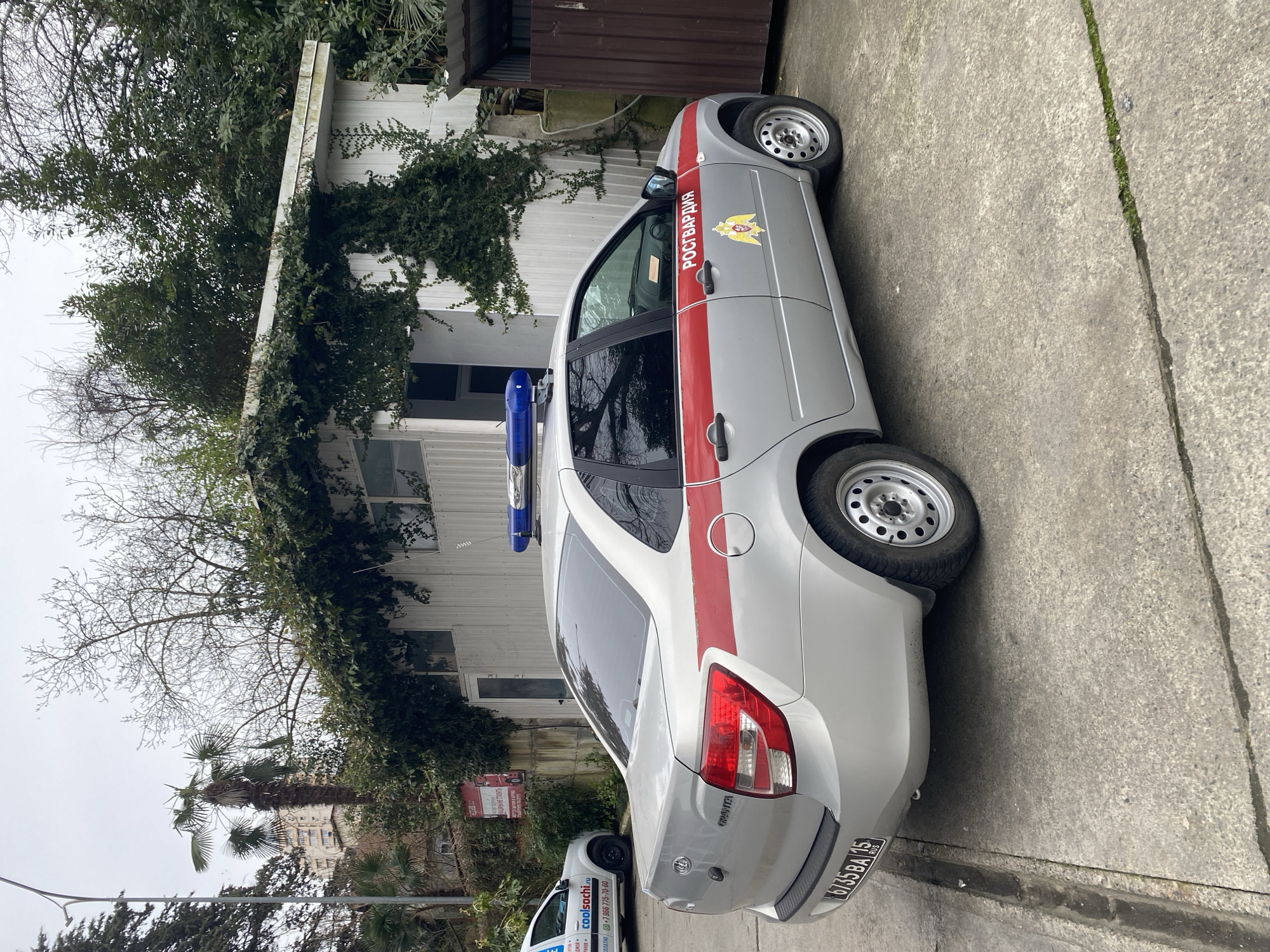 The image shows a parked vehicle, likely in the city of Sochi, Russia. The car has the markings and insignia of the Russian National Guard (Rosgvardia), visible by the "РОСГВАРДИЯ" text and emblem. A red stripe runs along the side of the vehicle. Blue emergency lights are mounted on the roof. The license plate reads "6735 BA 23 RUS." The car model is "GRANTA" (labeled in the back).
Behind the vehicle is a building with white siding. Vines and vegetation are covering part of the building's exterior. A second vehicle, partially obscured, is visible in the background. In the background, there are palm trees and what appears to be a building of significant size or height.