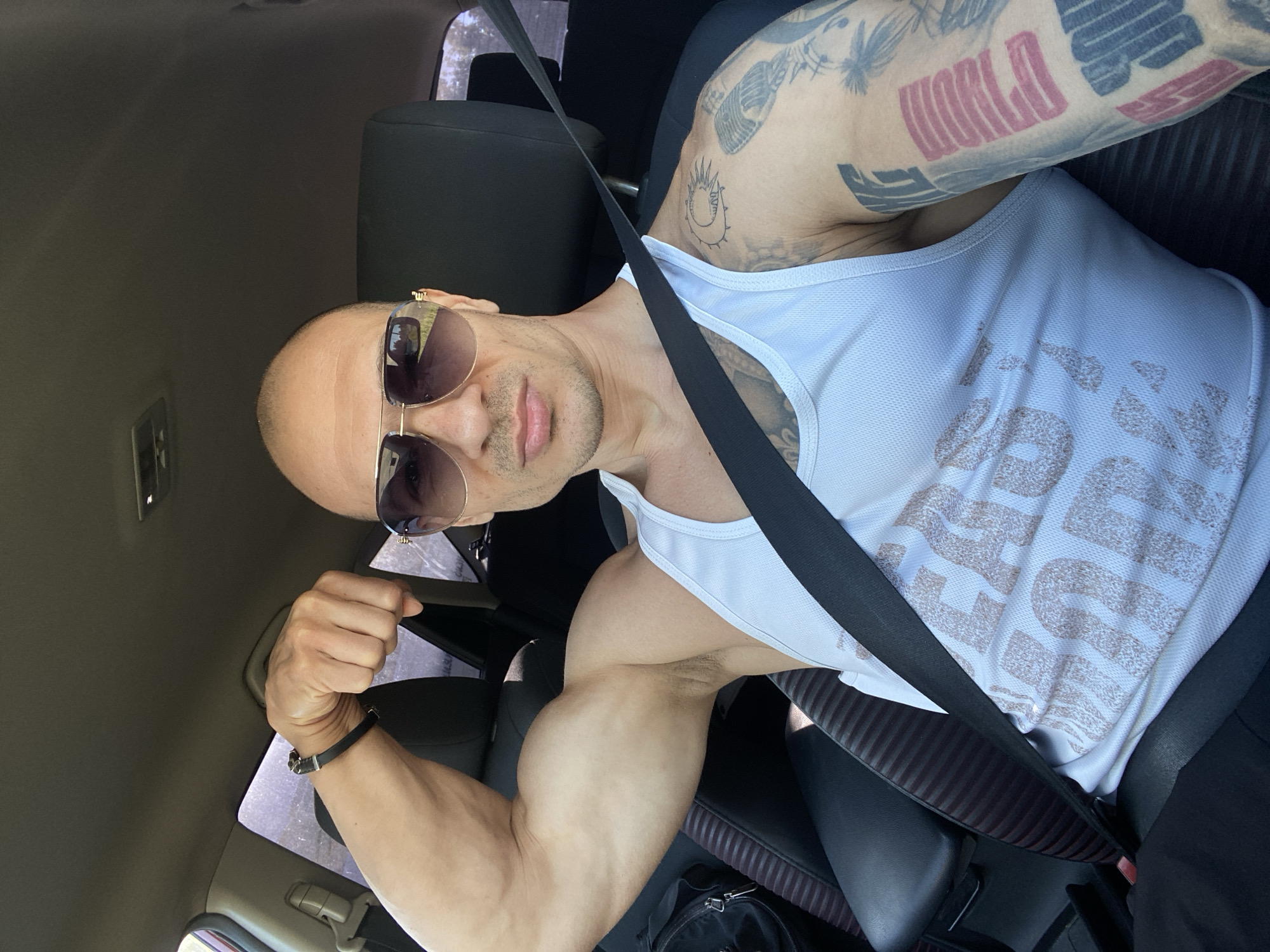A man wearing a white tank top and sunglasses is sitting in the driver's seat of a car. He is wearing a seatbelt. He is flexing his arm and has tattoos on both arms. The location is in a car, not specifically Pleven, Bulgaria.