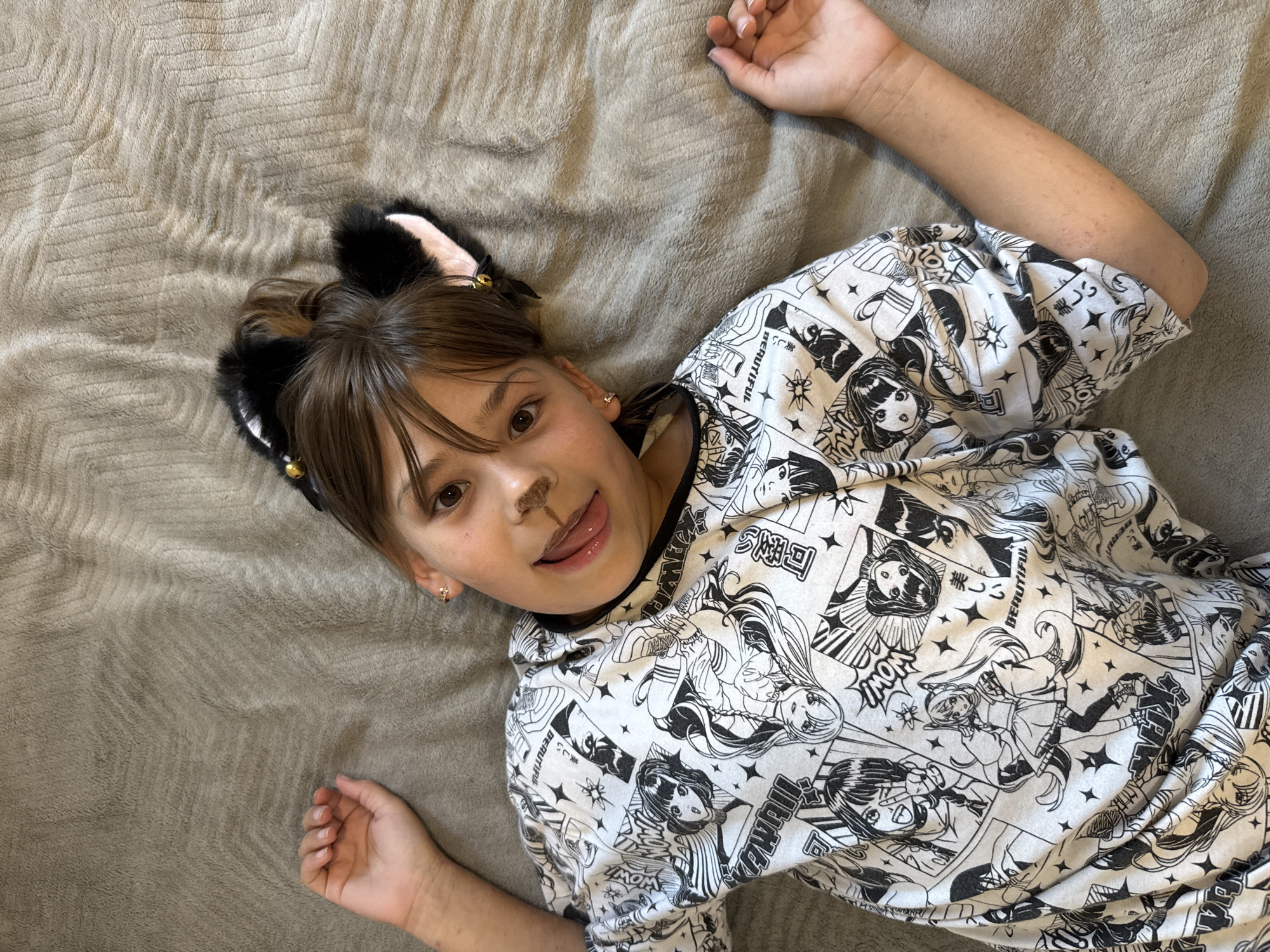 This media file shows a young person lying on a gray, textured blanket, looking up at the camera. They are wearing a black and white shirt with a graphic pattern of anime-style characters and text, including "BEAUTIFUL," "WOW!", and what appear to be Japanese characters. The person has on fuzzy black cat ears with pink insides and a small gold earring. There is also a smudge of brown makeup on their nose, resembling whiskers, and their tongue is sticking out slightly. Their right arm is extended upwards, and their left hand is resting on their lap. The lighting suggests it is daytime, possibly indoors. The background is a plain, textured blanket. Visible text includes "美しい," "WOW," "BEAUTIFUL," "WOW!", "可愛い," and "TIUME." The image's metadata indicates a location context of Rusanovo, Russia.