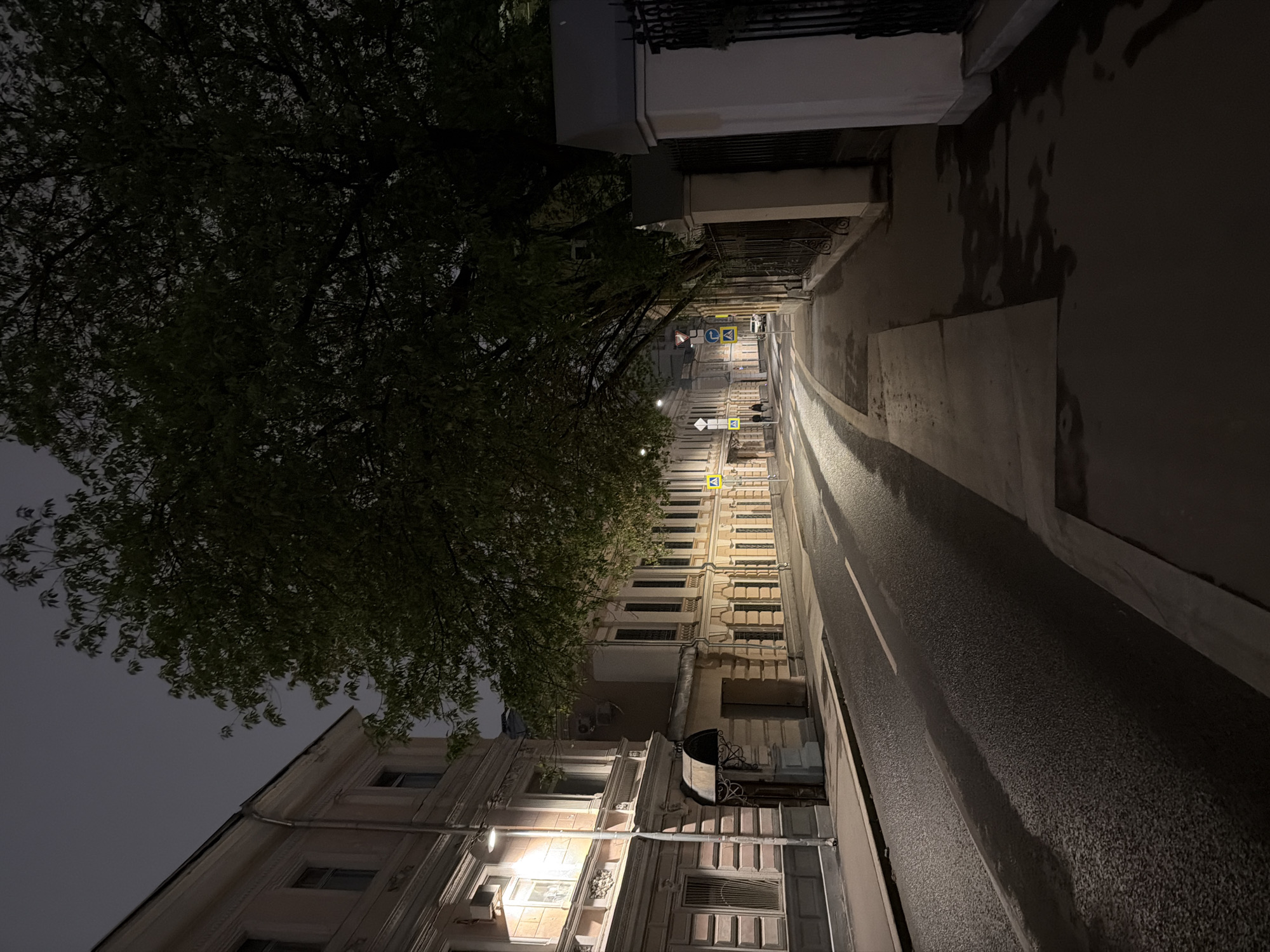 The image shows a dimly lit street in Moscow, Russia, at night. The street is narrow and lined with buildings on both sides. The buildings are primarily multi-story with architectural details like molding and decorative window frames. A large tree overhangs the street on the left. The street itself is asphalt and appears wet, reflecting the light from street lamps and building windows. Several road signs are visible in the distance, indicating the street is open to traffic. There are a few indistinct figures walking further down the street. The overall scene has a quiet, urban atmosphere.