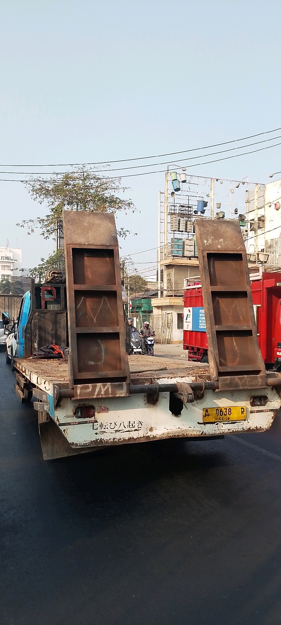 In an outdoor, urban setting in Bekasi, Indonesia, a tow truck with a flatbed trailer is parked on a paved road. The truck is white with blue accents and has its rear ramp lowered. Two large, rusty metal ramps are propped up at the back of the trailer, forming a channel. The trailer bed is stained and weathered. A yellow license plate with the characters "A 9638 P" is visible. White lettering, possibly in Japanese or Chinese, is visible on the rear of the trailer below the license plate. A red truck is parked to the right of the tow truck. In the background, there are buildings, trees with sparse foliage, and power lines. Two individuals on a motorcycle are passing by. The sky is clear and pale blue.